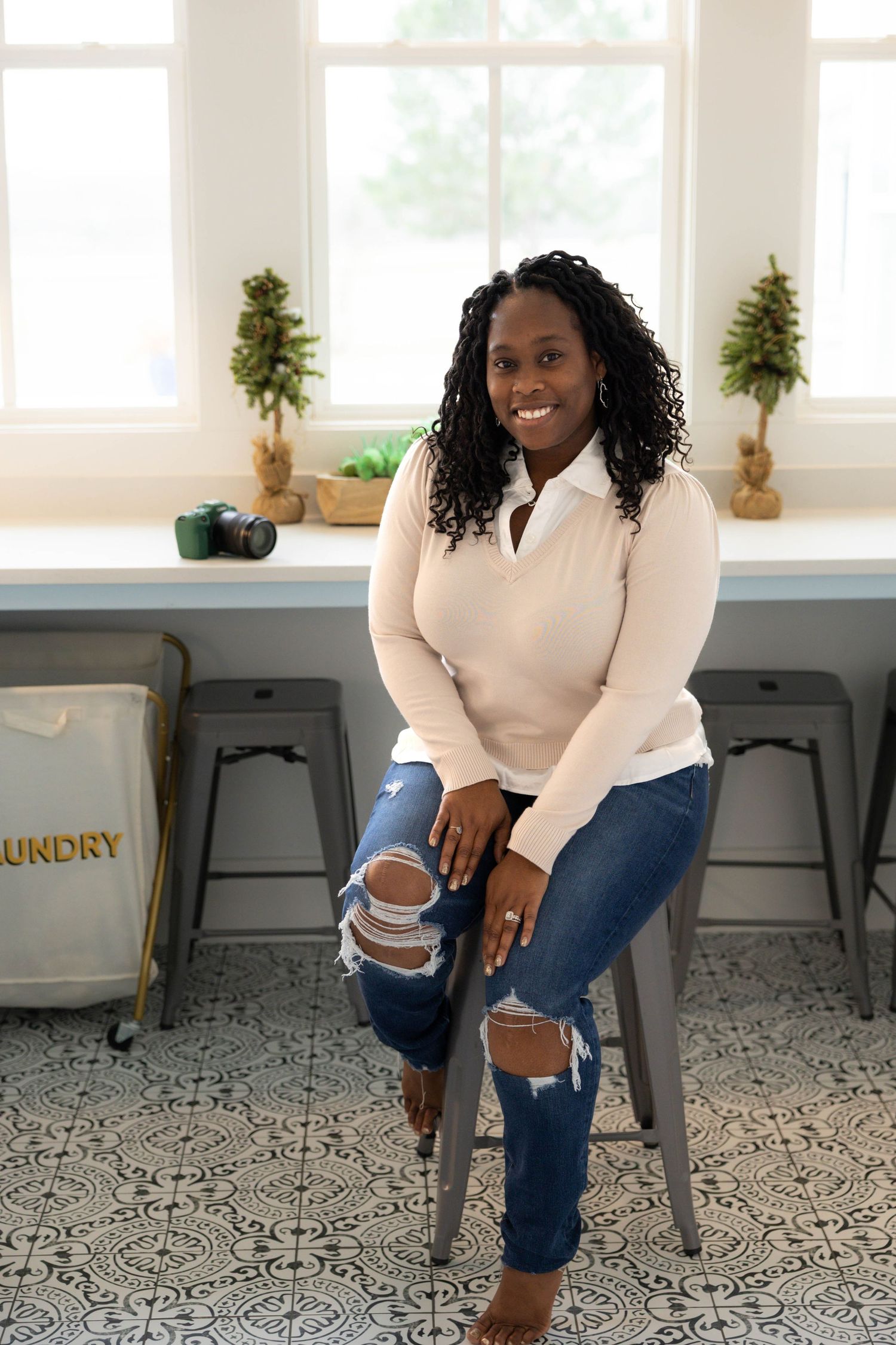 Person in white sweater and distressed jeans sits on gray chair against white wall with window decor.