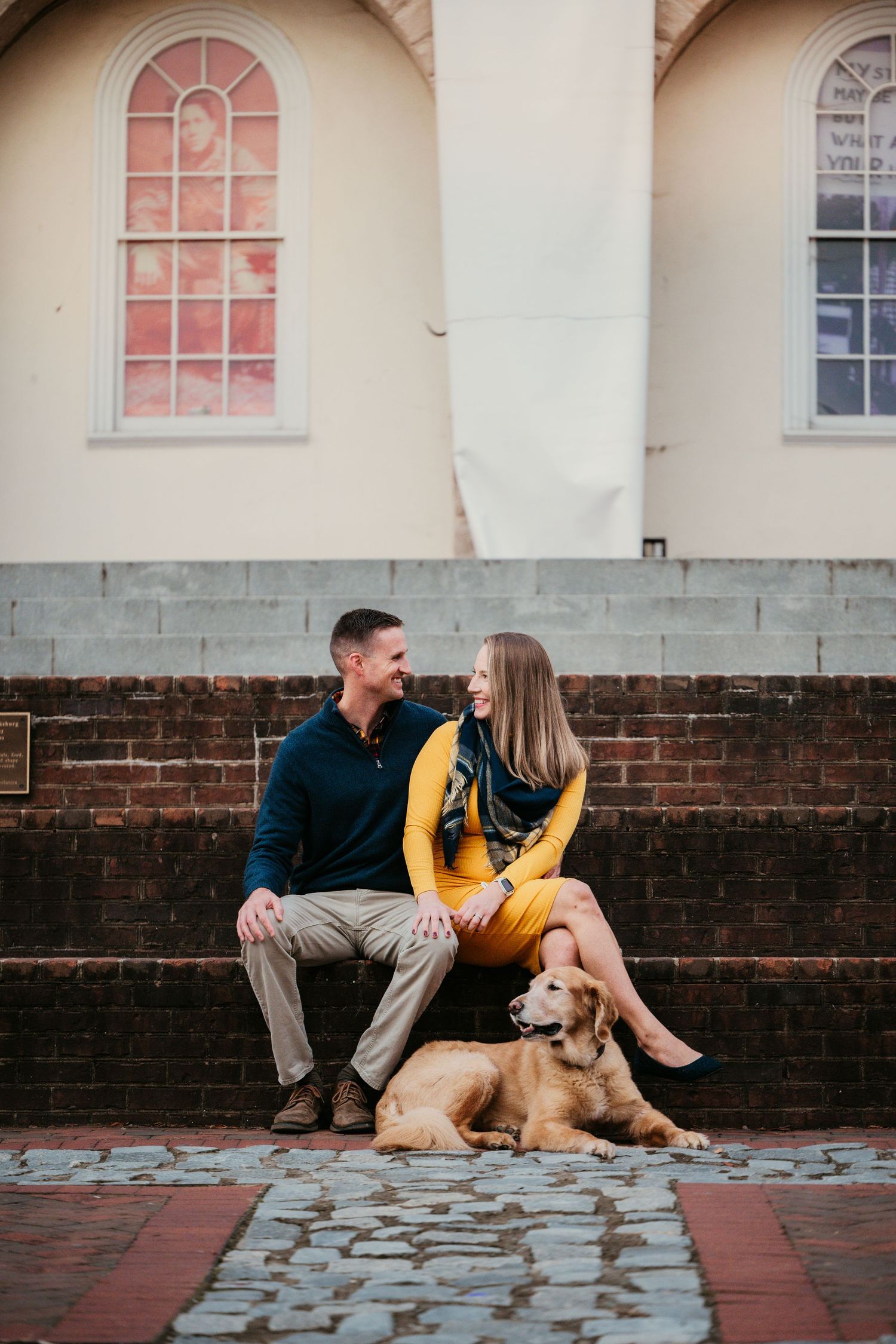 Family relaxing on brick steps with dog against historic building with arched windows.