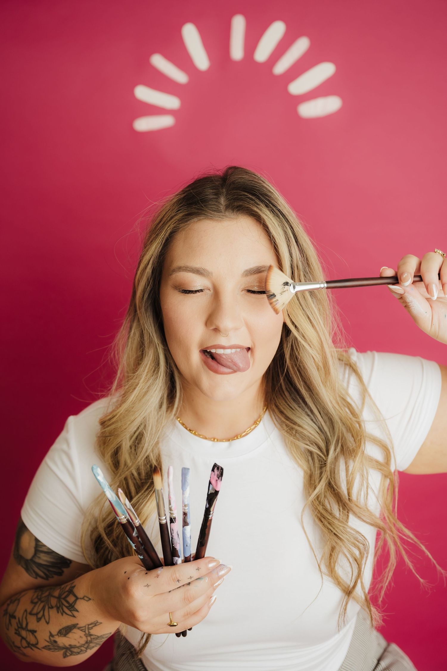 Blonde woman with tattoos holds makeup brushes, playfully applying one to her eye against a pink background.