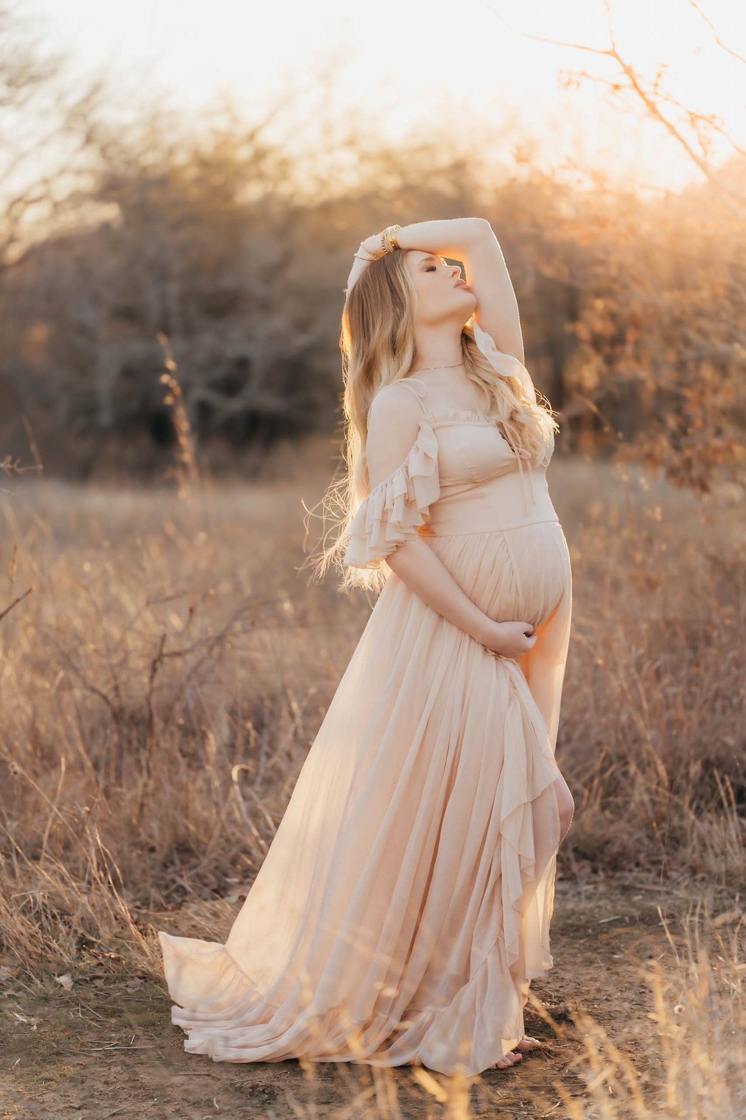 Pregnant woman in blush gown posing at golden hour in a field, cradling her baby bump with flowing hair.