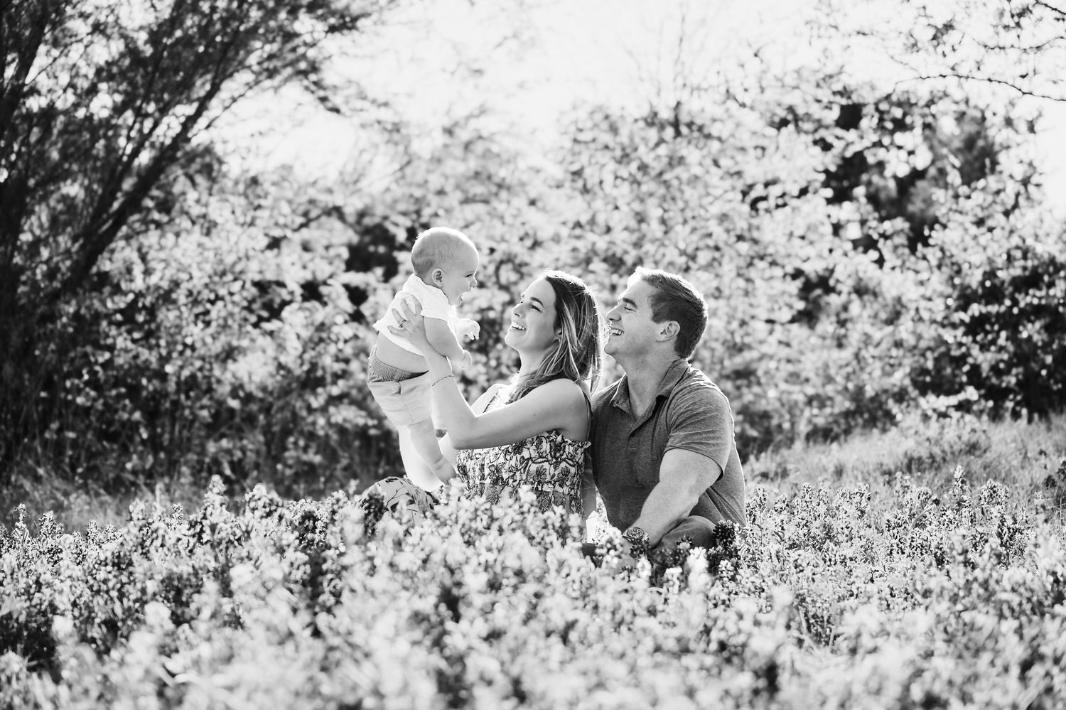Black and white photo of a couple lifting their baby in a sunlit meadow surrounded by  bluebonnets.  Northlake Tx, Knot & Wing Photography