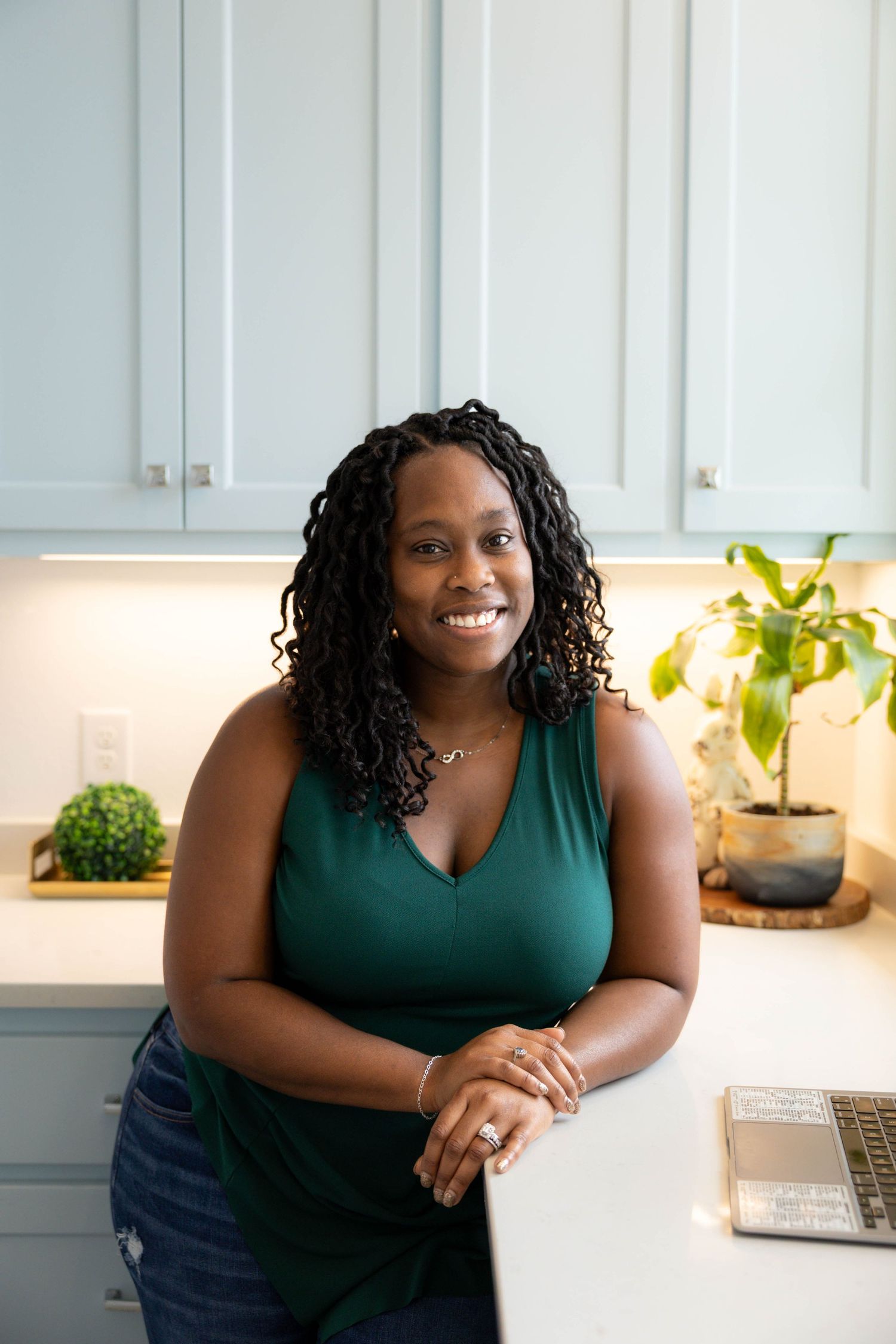 Person in a green sleeveless top smiling while standing in a modern kitchen with white cabinets and plants.