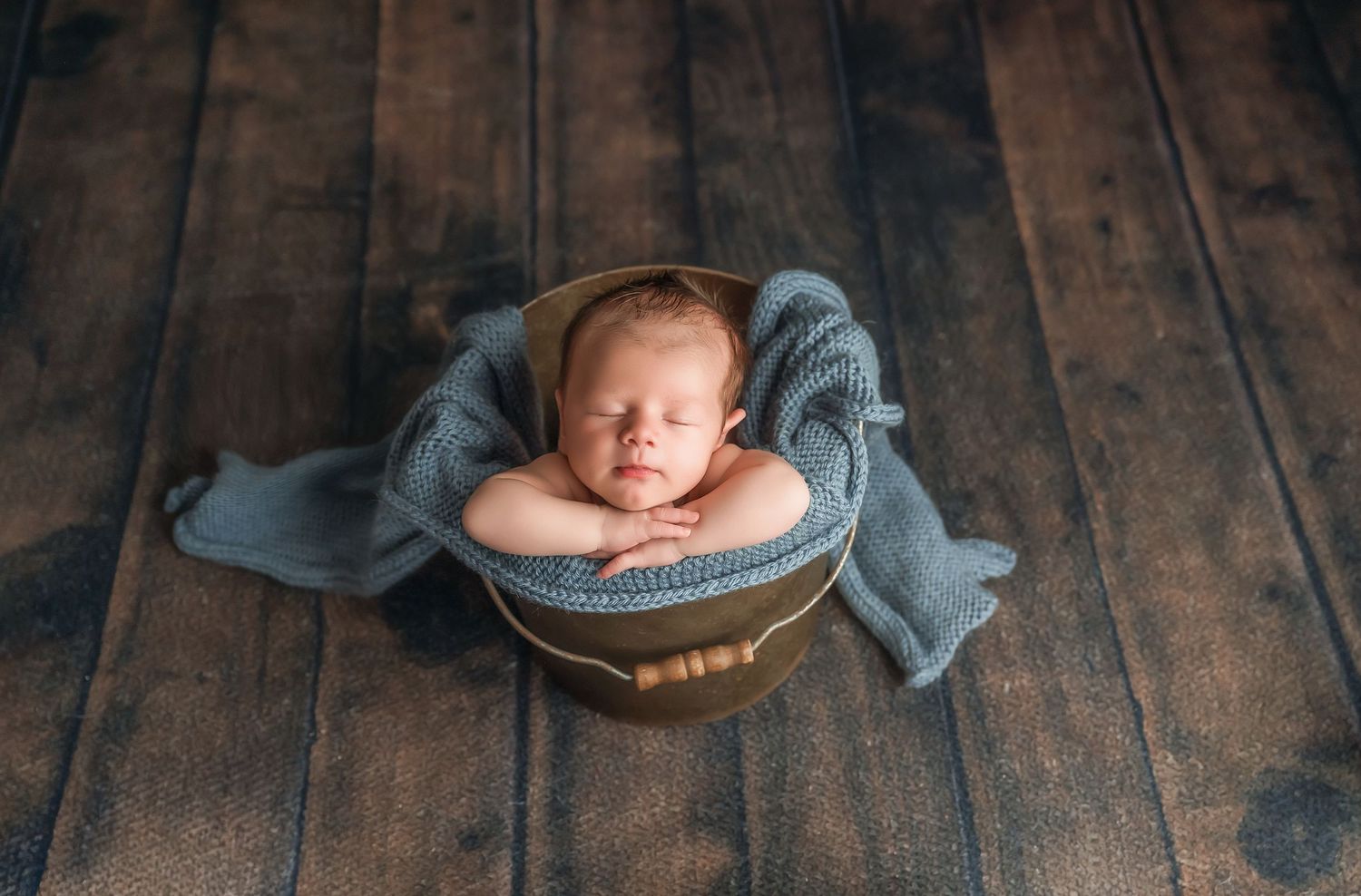 Newborn baby sleeping peacefully in a small bucket wrapped in a soft blue knit blanket on a dark wood floor.
