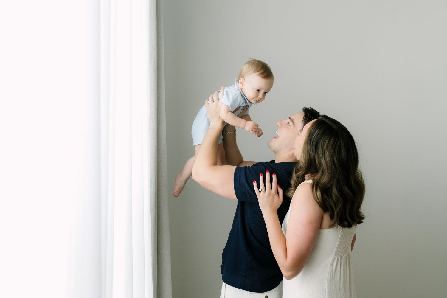 A joyful mother lifts her smiling baby in the air near a bright window in a light-filled studio setting.