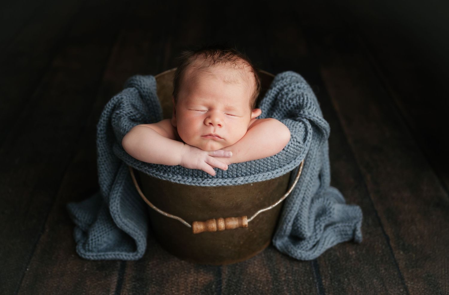 Sleeping newborn wrapped in soft gray blanket rests peacefully in vintage metal bucket on wooden floor.