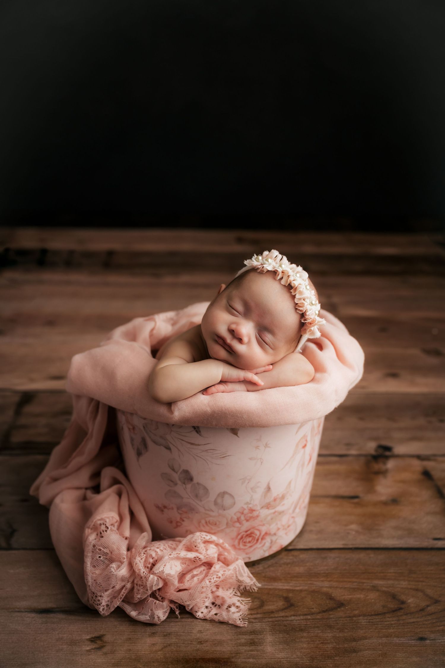 A sleeping newborn rests peacefully in a pink vintage container on rustic wooden floorboards.