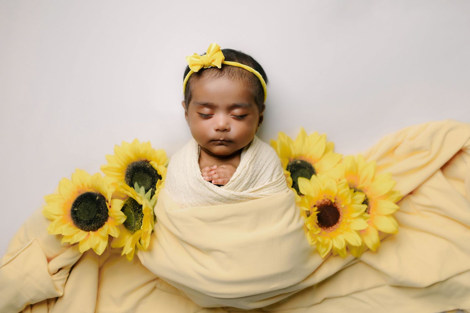 Newborn baby girl swaddled in yellow wrap with sunflowers and yellow headband on soft yellow blanket.