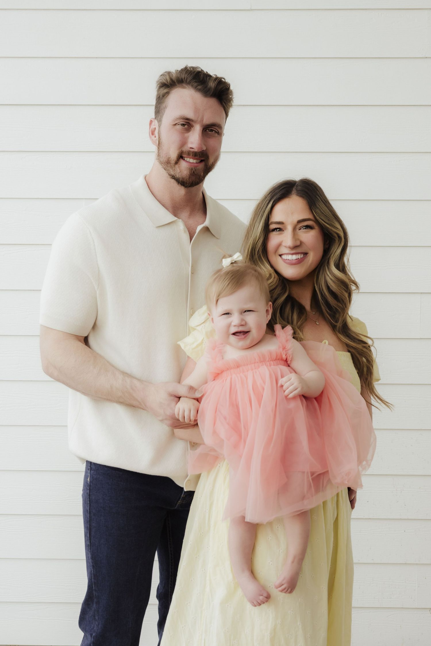 A smiling family of three poses together, with parents holding their laughing baby girl in a pink tulle dress.