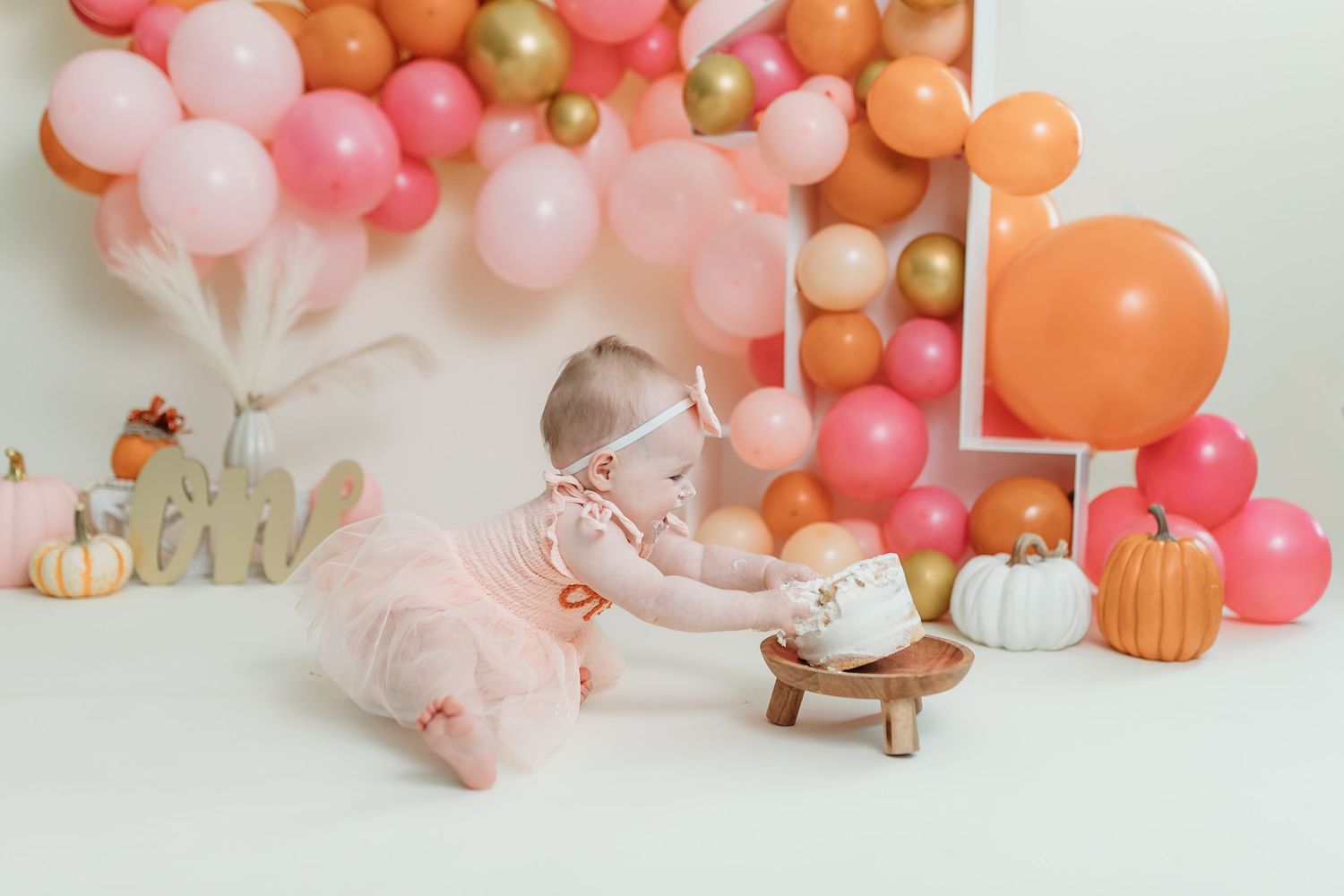 Festive balloon arch installation in coral, peach, and gold tones with pumpkin decorations and fall-themed props.