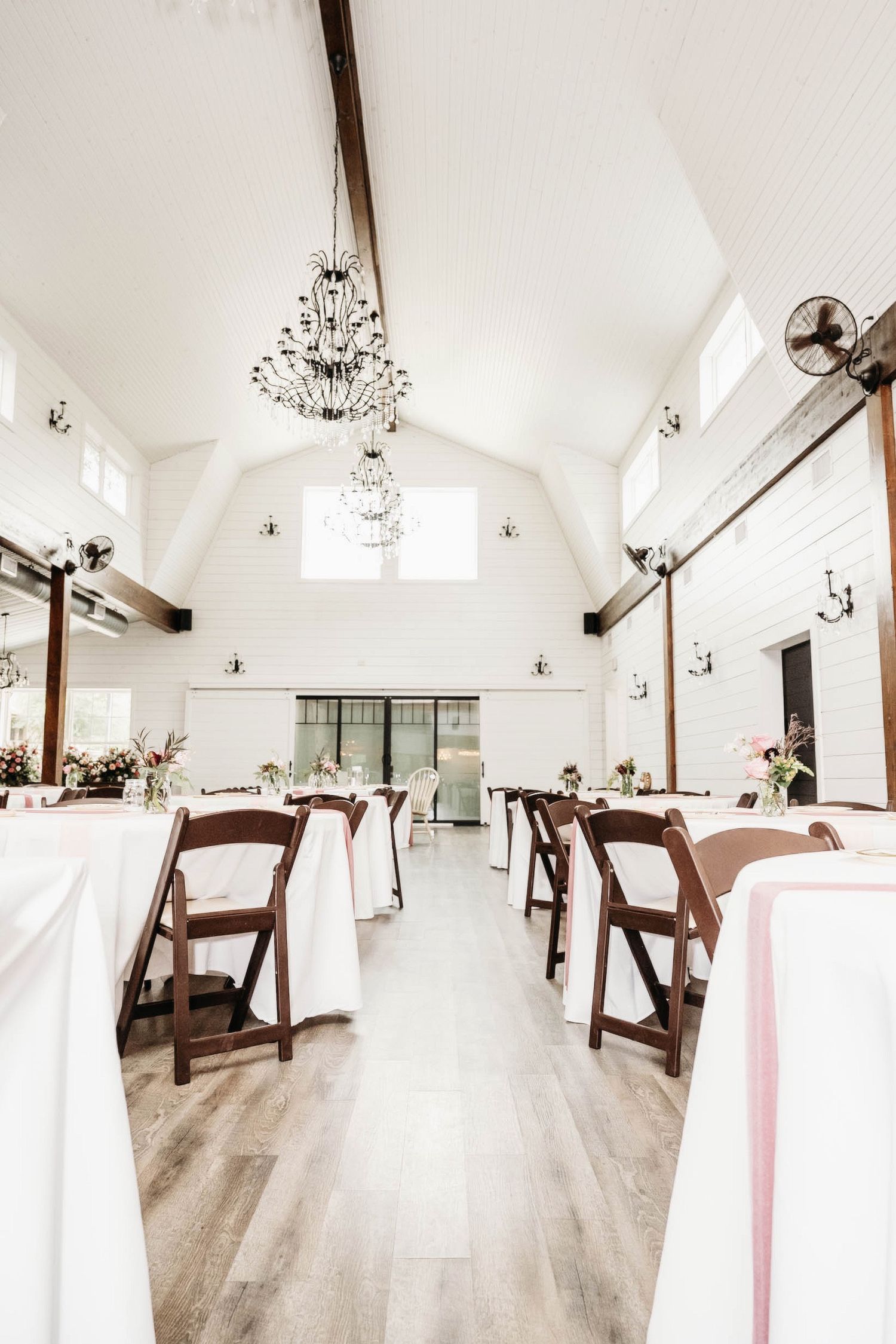 Elegant white wedding venue interior with crystal chandeliers, wooden beams, and folding chairs arranged around tables.