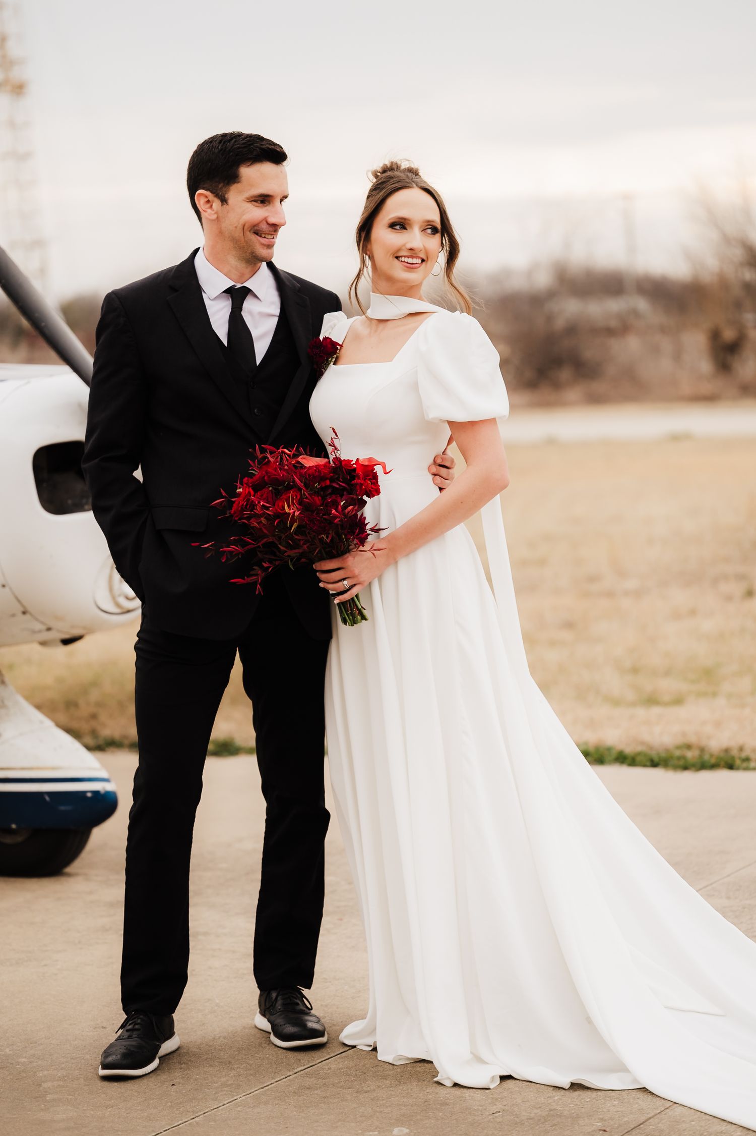Wedding couple in elegant attire poses outdoors, with the bride in white and groom in black holding deep red flowers.