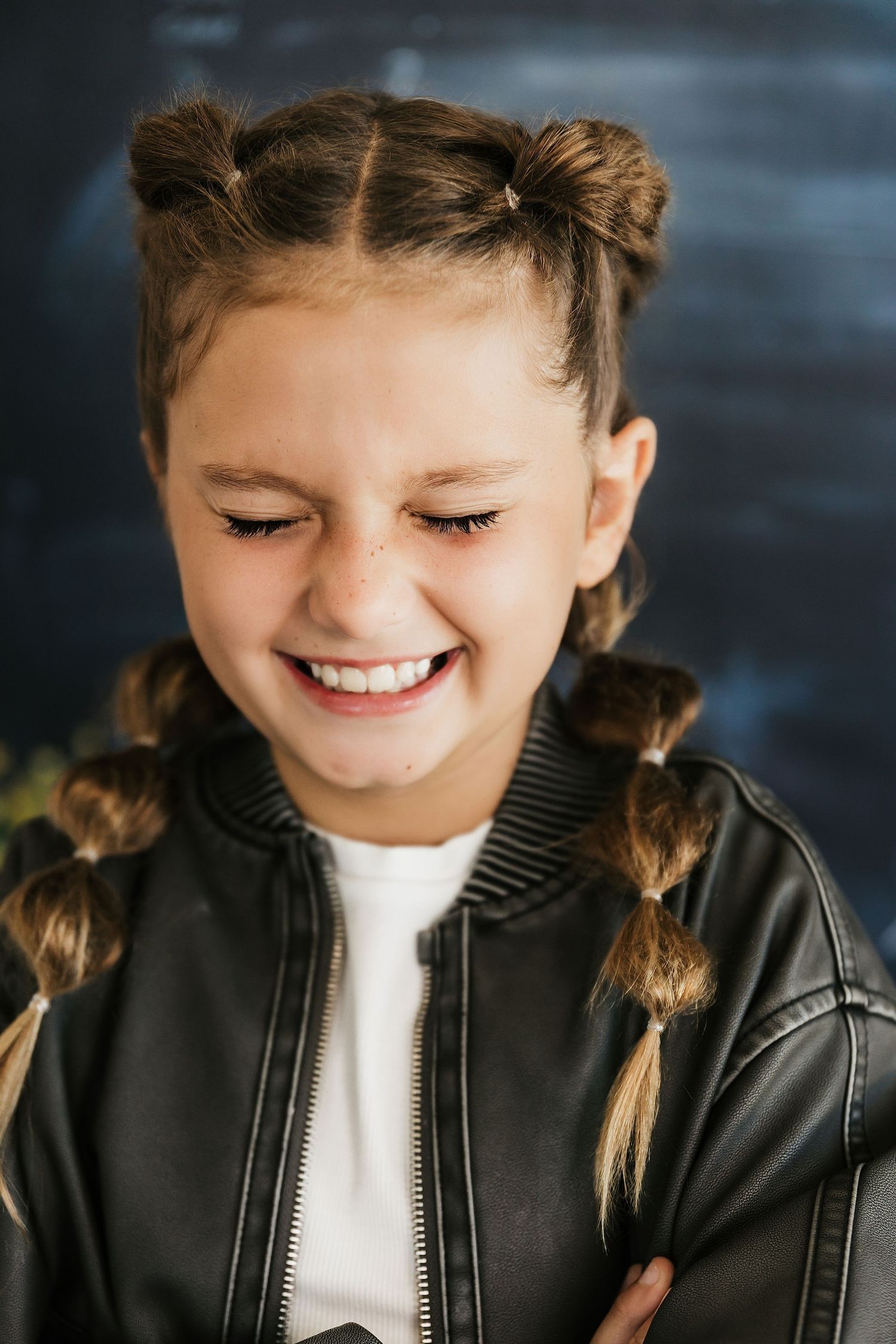 A young girl with braided pigtails and a black leather jacket smiles brightly against a dark background.