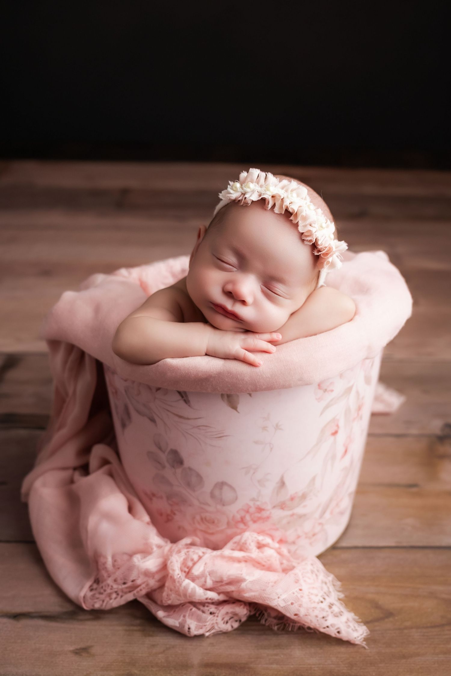 Sleeping newborn baby girl in pink floral bucket with lace wrap and flower headband on wooden floor.