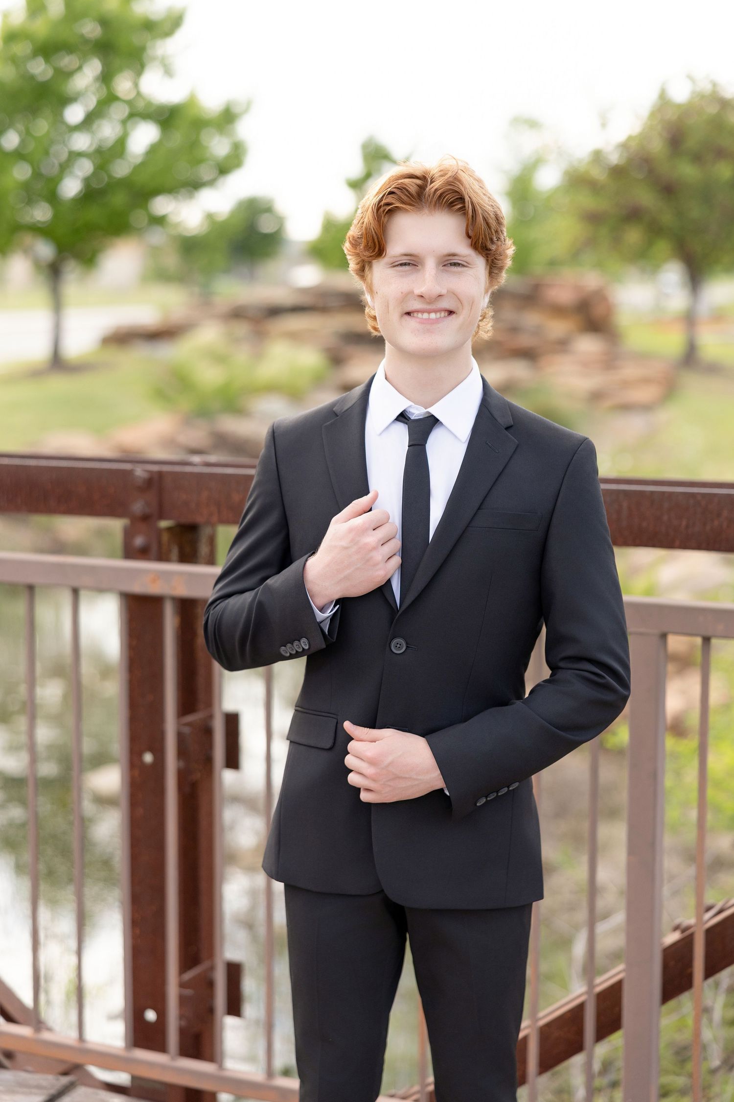 Smiling young man in black suit and tie stands on bridge in outdoor park setting.