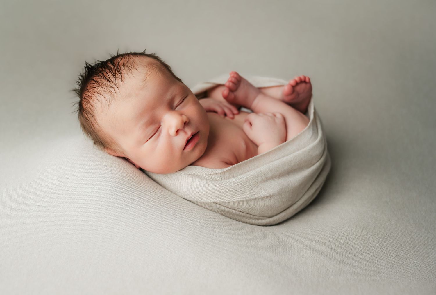 Sleeping newborn baby wrapped in soft white fabric against neutral gray background.