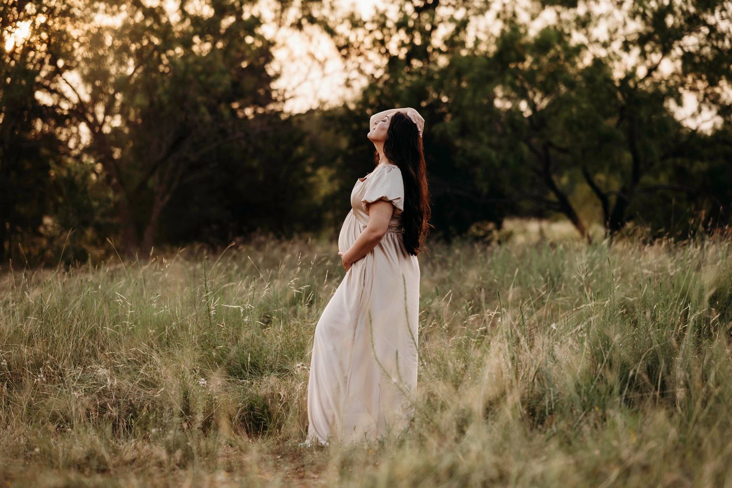 A long flowing white dress catches the golden sunset light in a grassy meadow field during a photo session.