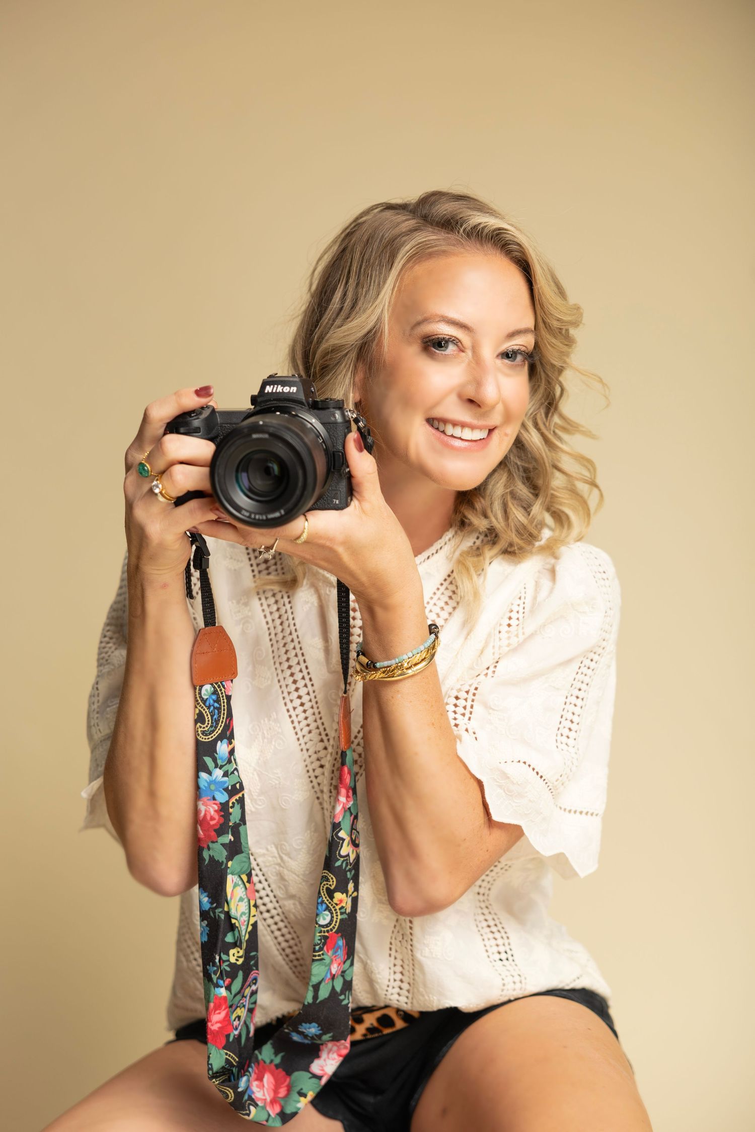 Professional photographer with blonde hair holds camera while posing in white eyelet top and floral camera strap.