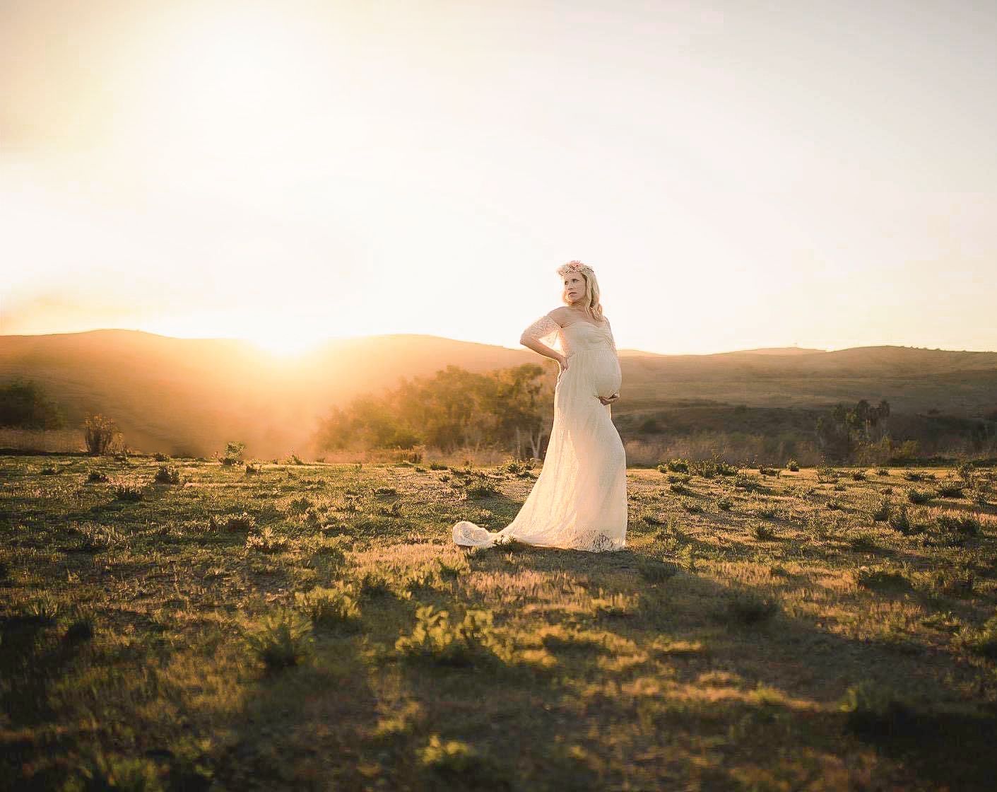 A bride in a flowing white dress stands silhouetted against a golden desert sunset in a panoramic landscape.