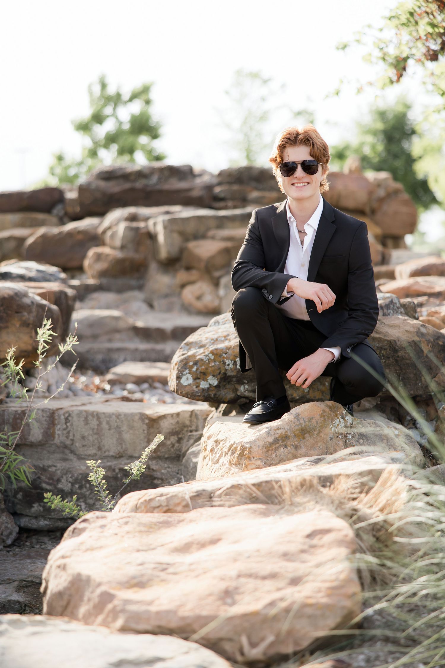 Young man in black suit and sunglasses sits on rocky stone steps smiling outdoors.