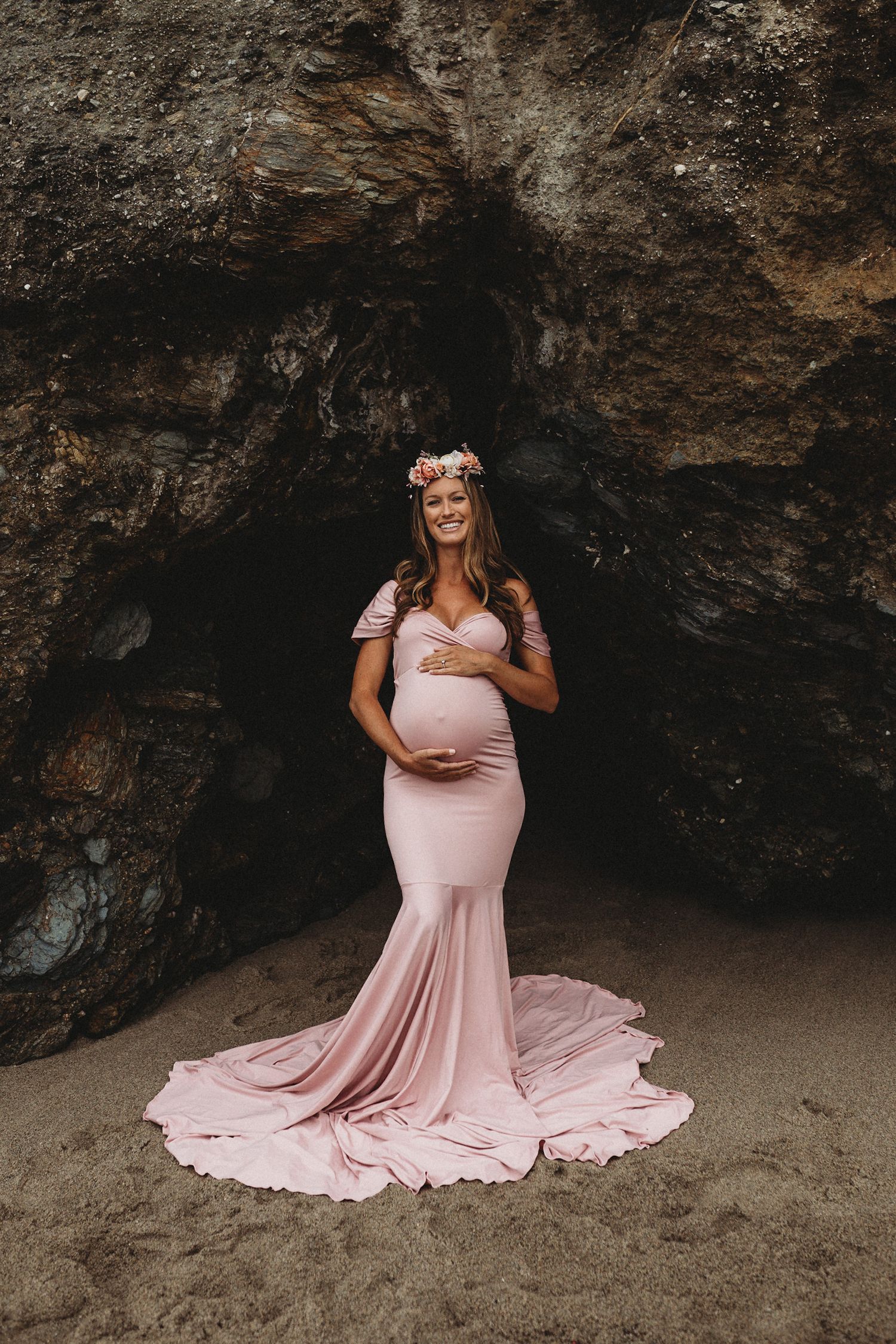 Maternity photography session featuring flowing pink dress against dramatic rocky beach cave backdrop.