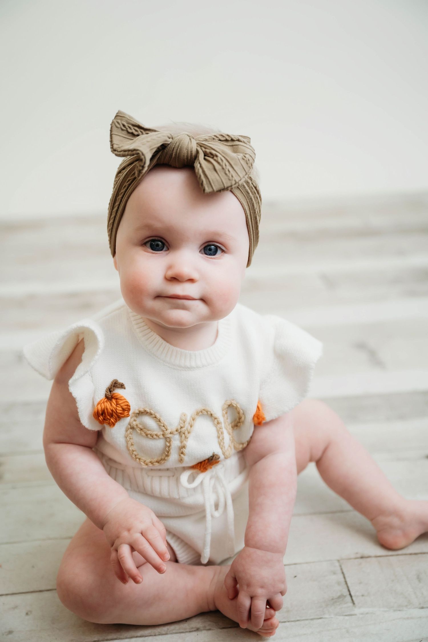 Baby wearing a knit headband and white outfit sits on light wooden floor showing progression of poses.