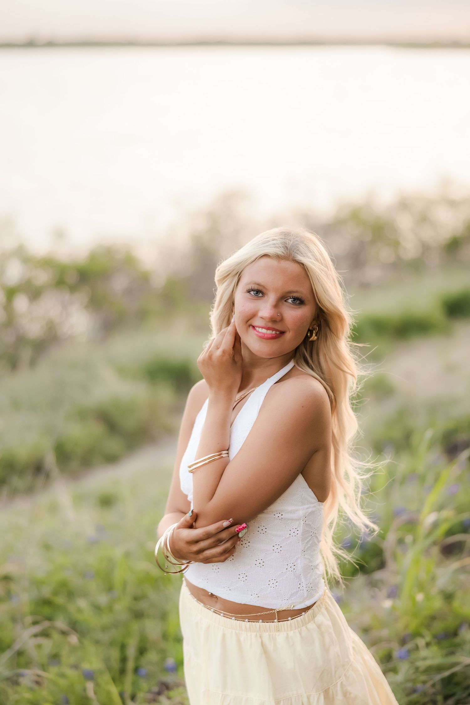Smiling blonde woman in white crop top and yellow skirt poses outdoors near water in golden hour light.