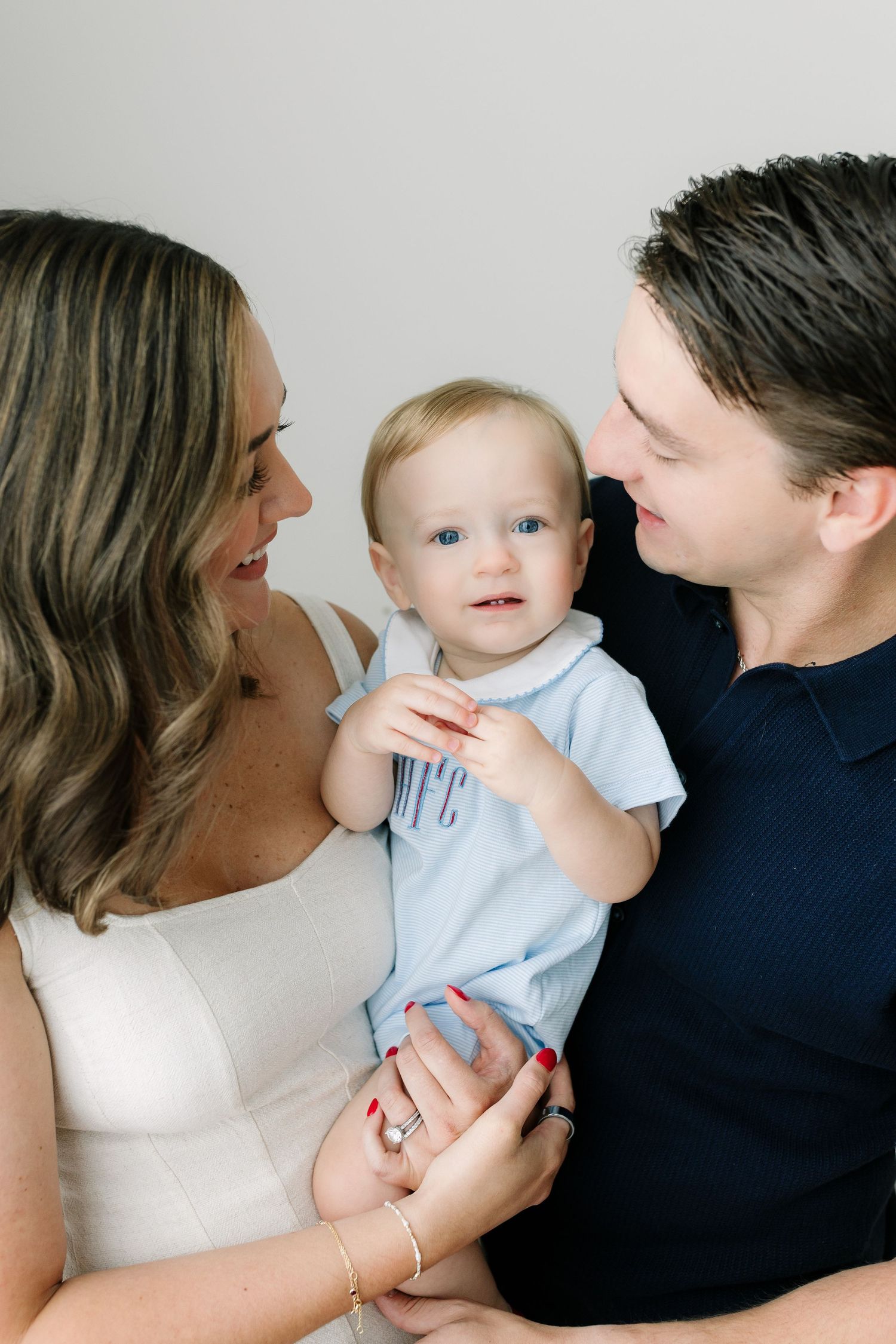 A smiling couple in a bright studio holds their blue-eyed baby boy dressed in a light blue outfit.