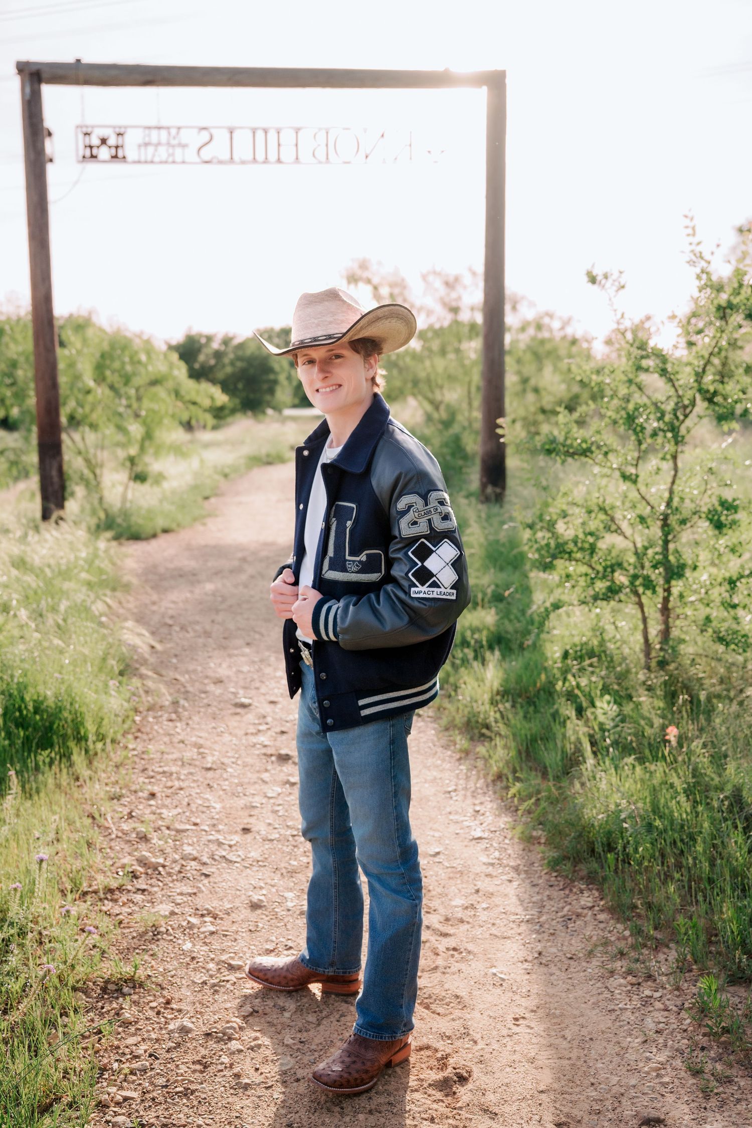 Young man in cowboy hat and letterman jacket poses on dirt path surrounded by greenery.