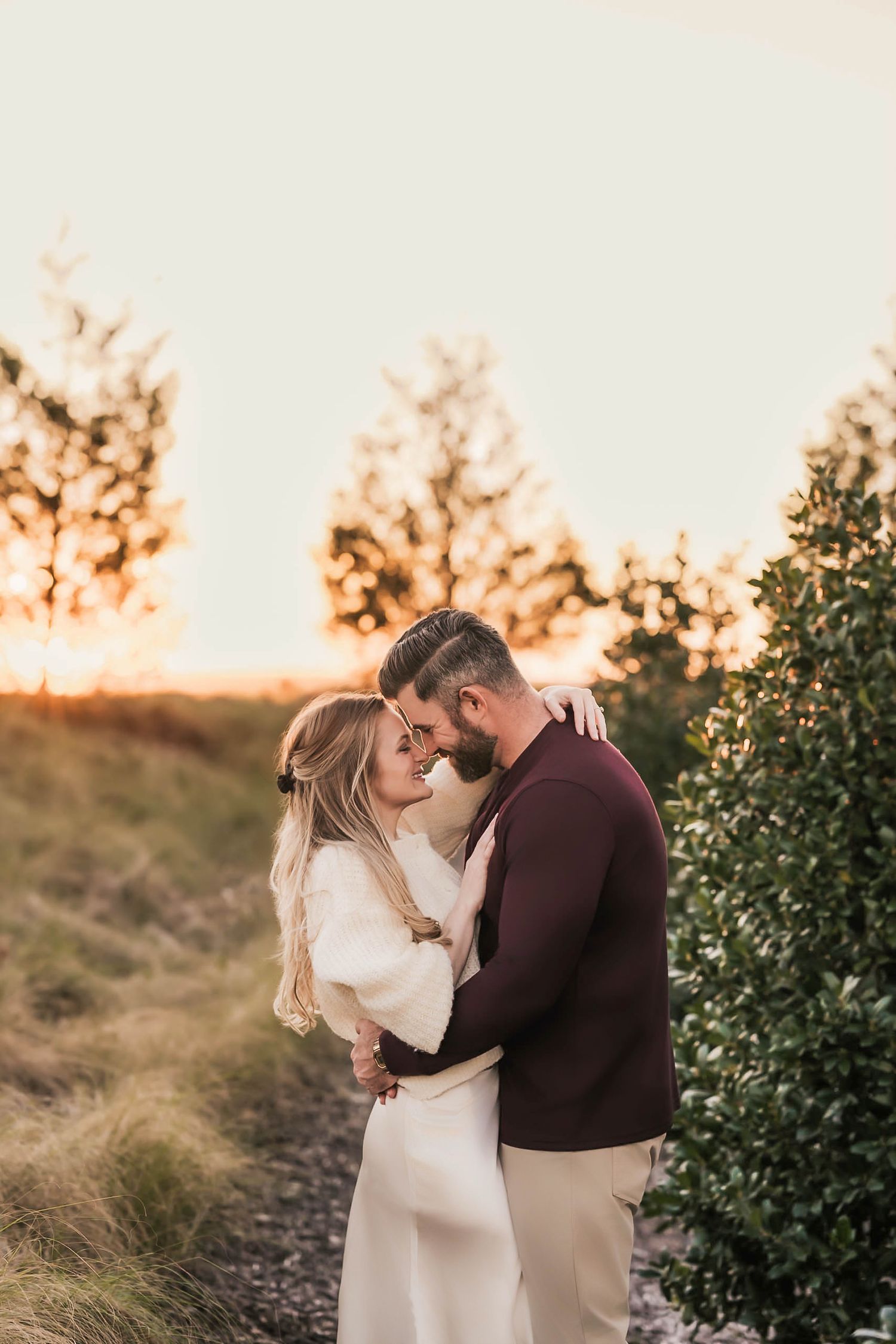 A romantic couple embraces in front of greenery during a golden sunset in a natural outdoor setting.