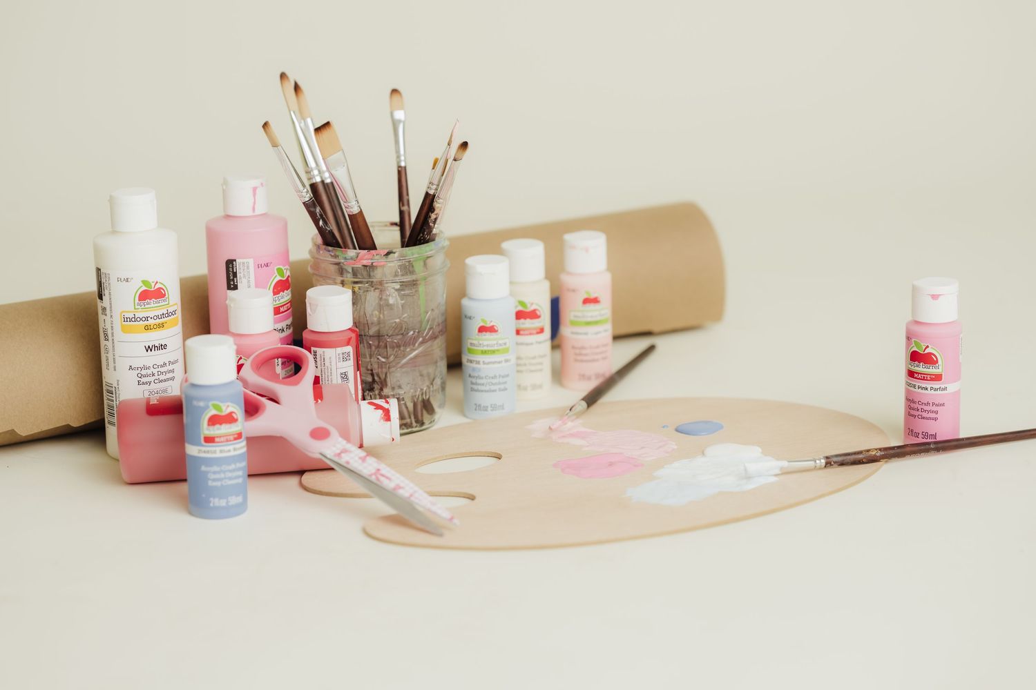 Acrylic paint bottles, brushes in a glass jar, and a wooden palette on a light beige surface for an art project.