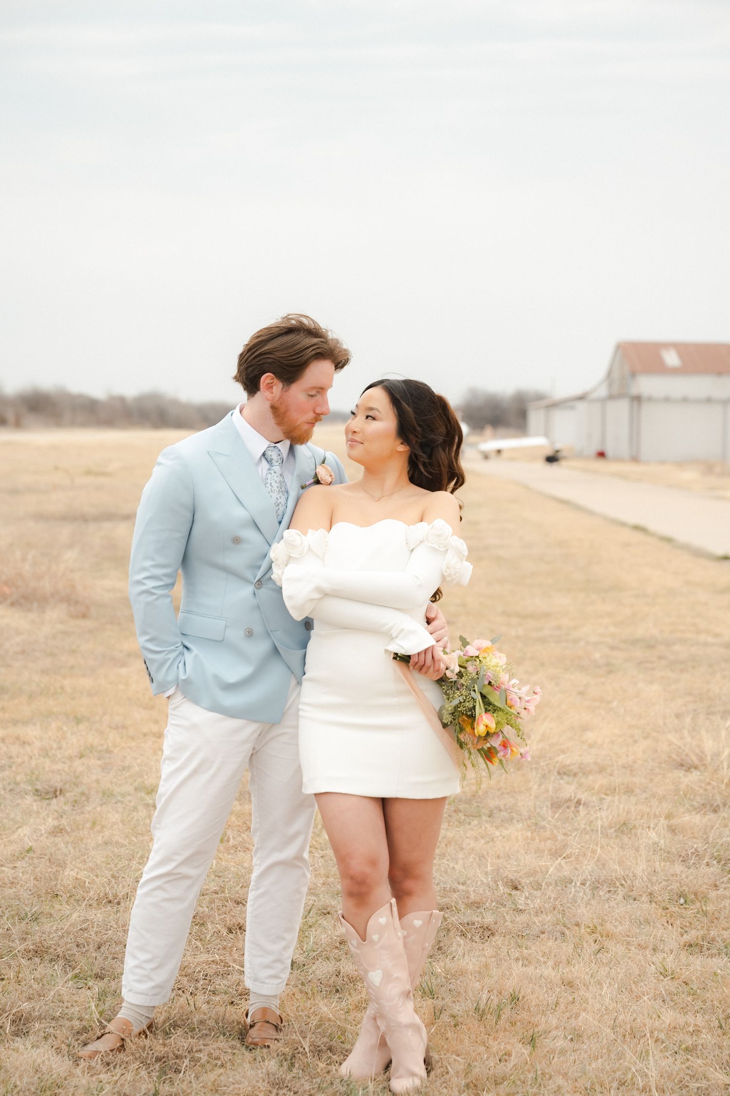 A romantic couple in light blue and white outfits embrace in a rustic field setting during a photo session.