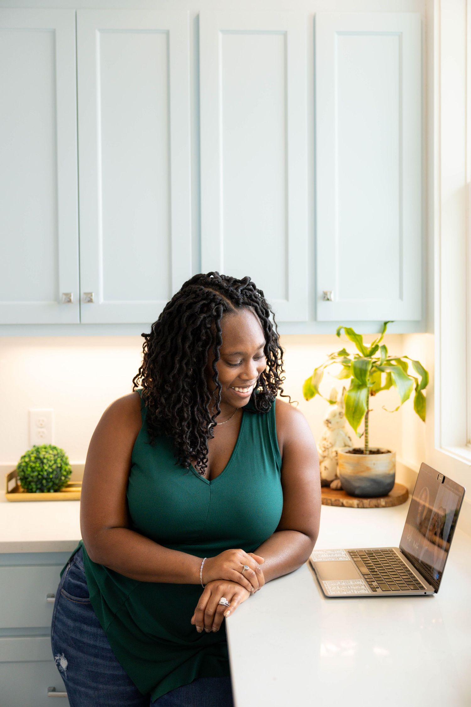 Person in teal sleeveless top works at white desk with laptop and plants in bright home office setting.