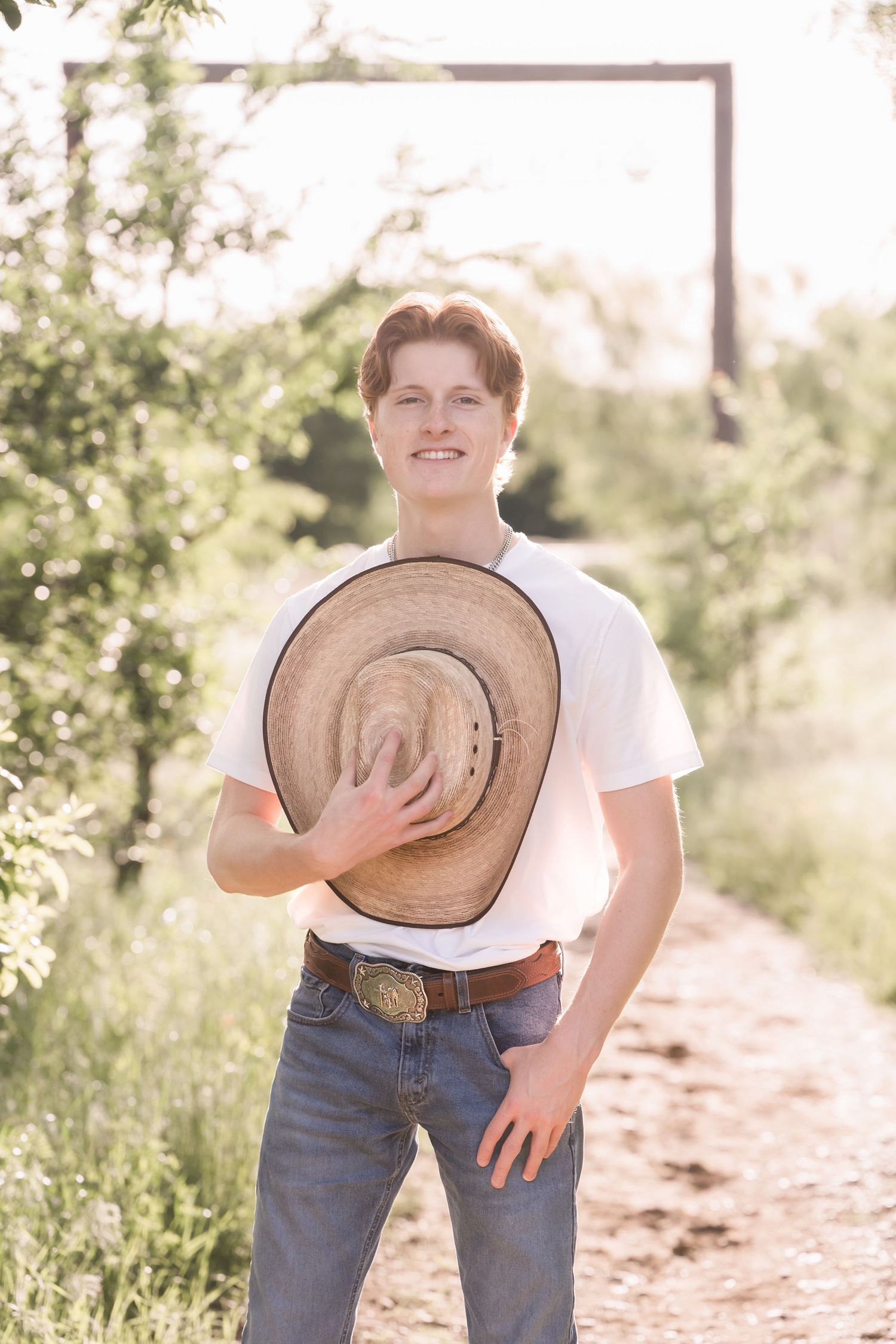 Smiling young man in white t-shirt and jeans holding a straw cowboy hat on a sunny rural dirt path.