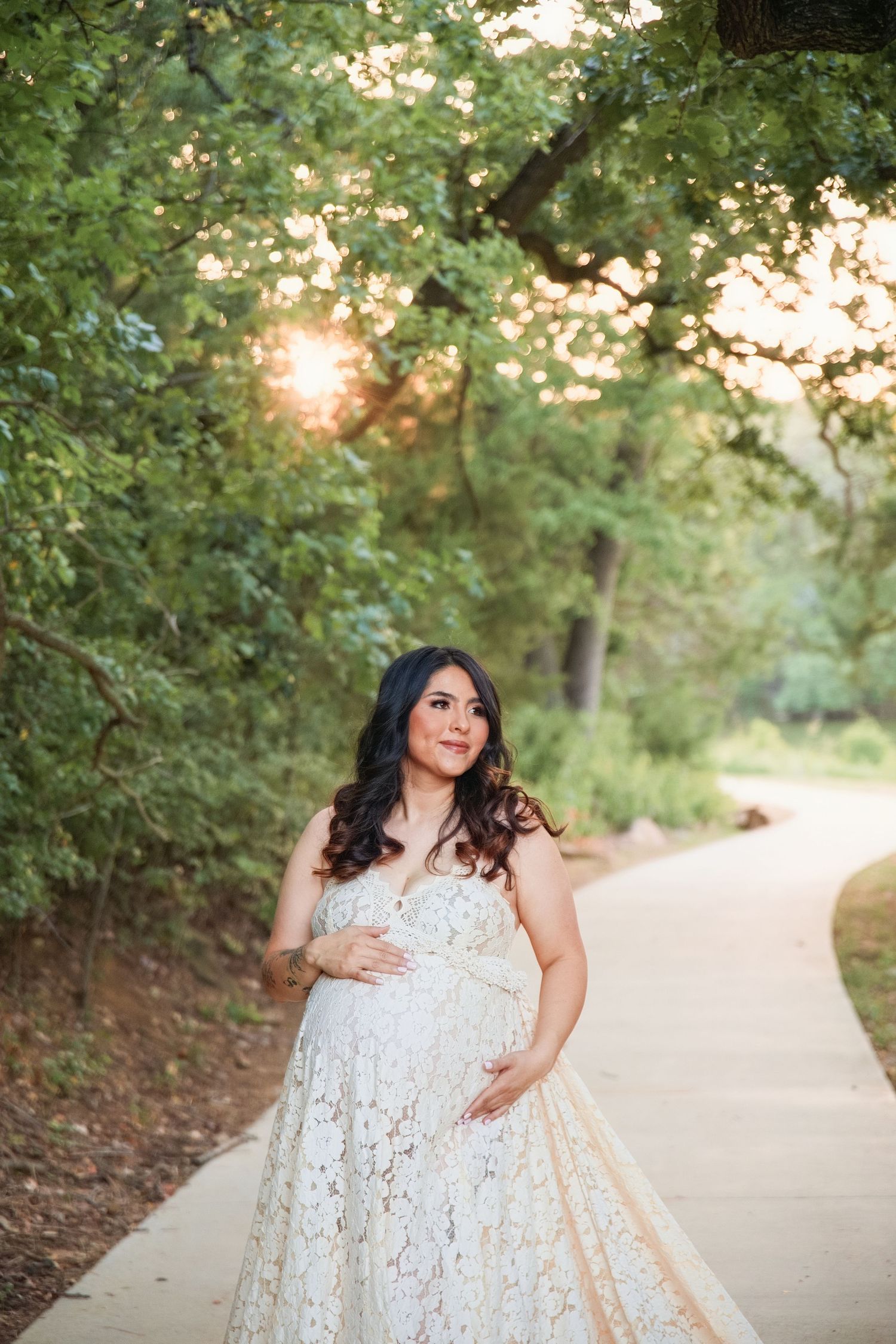 Pregnant woman in white lace gown cradling her belly on a sunlit park path surrounded by lush green trees.