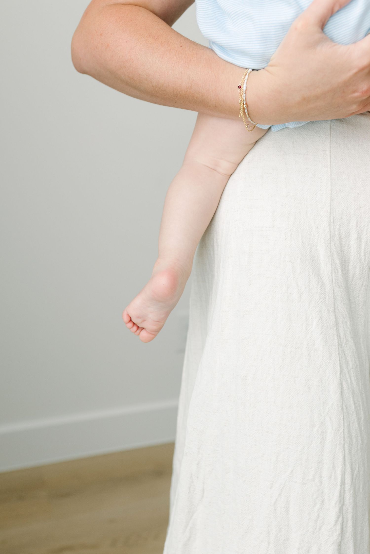A mother in a white linen skirt holds a baby, with the infant's tiny legs and feet visible against her outfit.