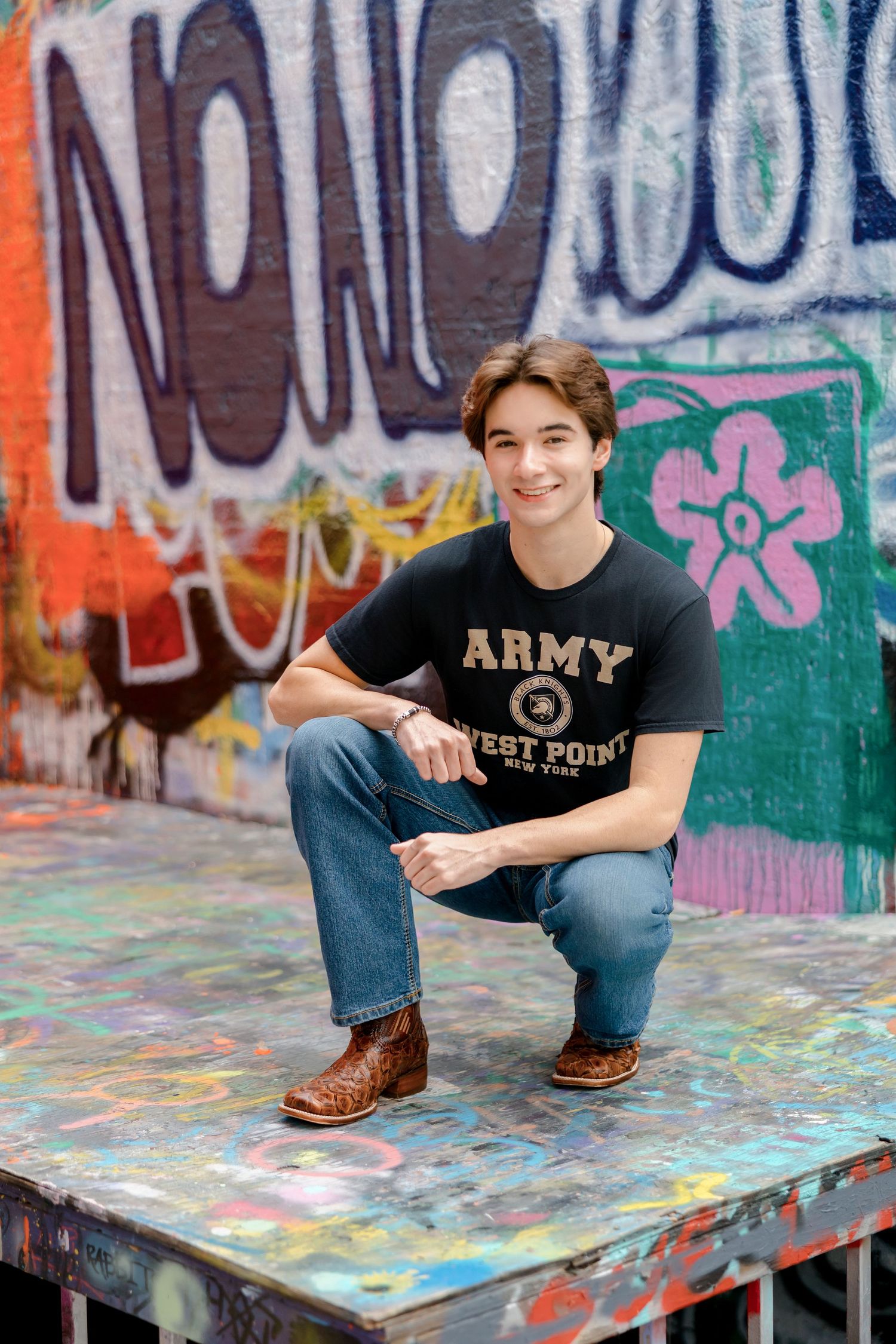 Young man in Army West Point shirt crouching on painted surface in front of colorful graffiti wall.