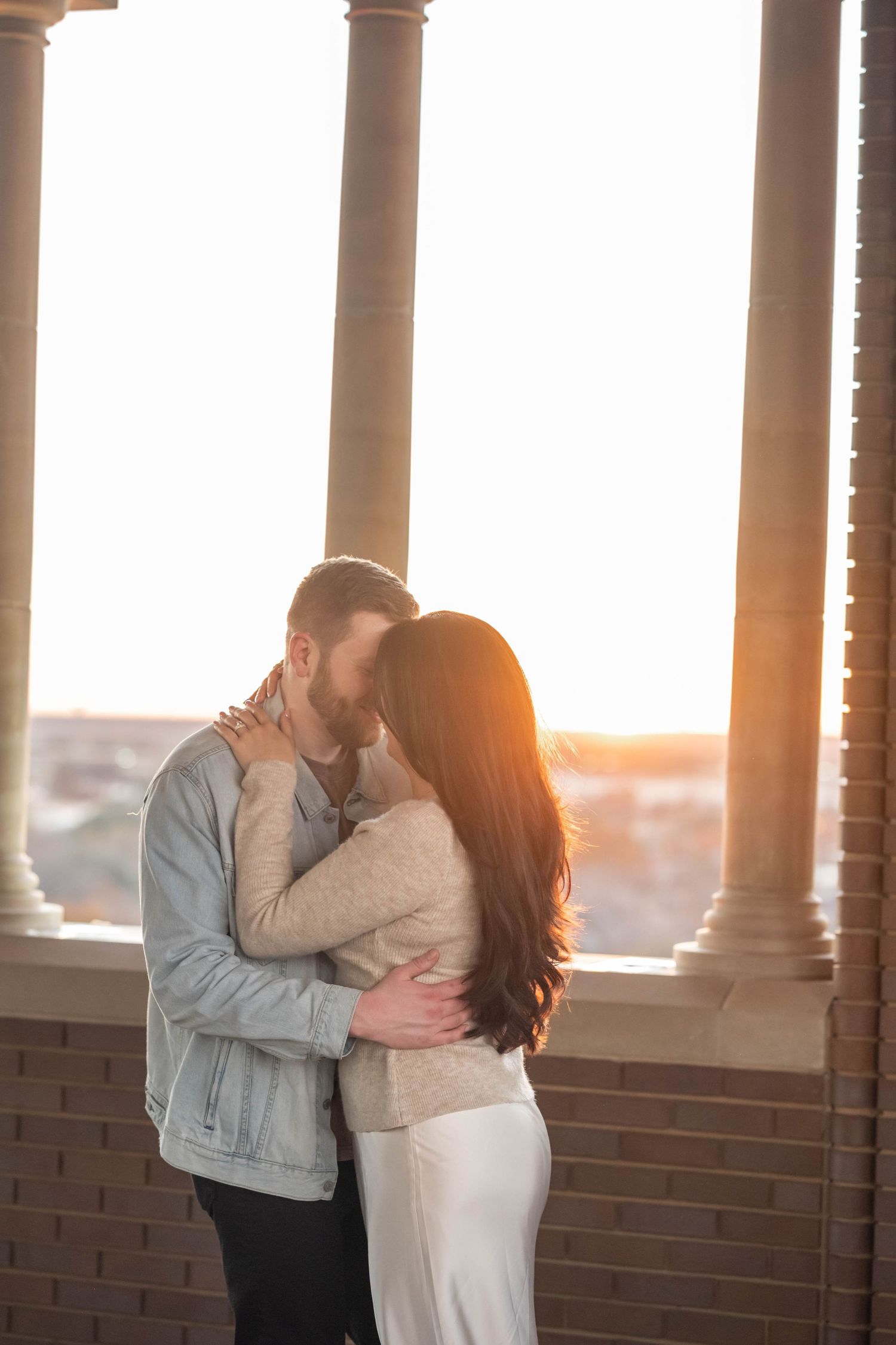 A couple embraces in a romantic moment at sunset, framed by elegant columns and brick walls by Knot & Wing Photography