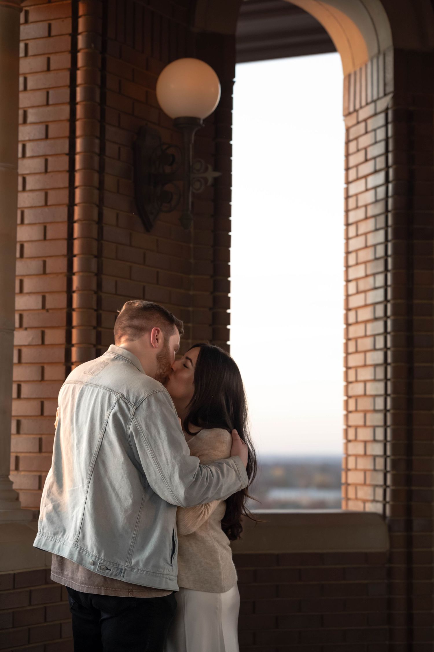 A romantic couple sharing an intimate moment in a brick archway at sunset with a glowing globe light fixture above.
