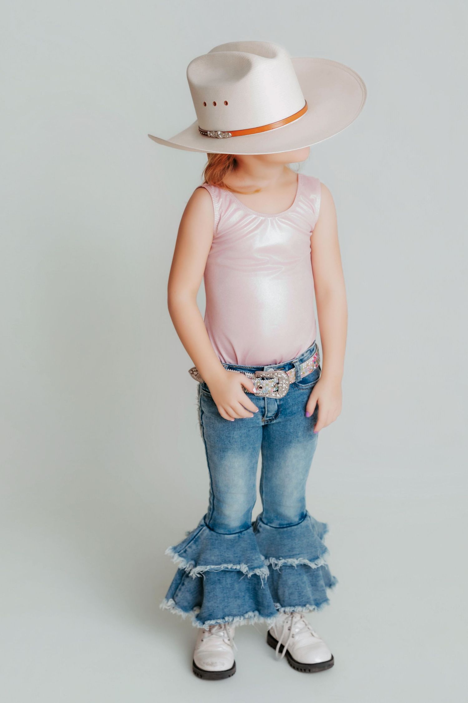 A toddler in a pink tank top, frayed denim jeans and cowboy hat stands against a white background.