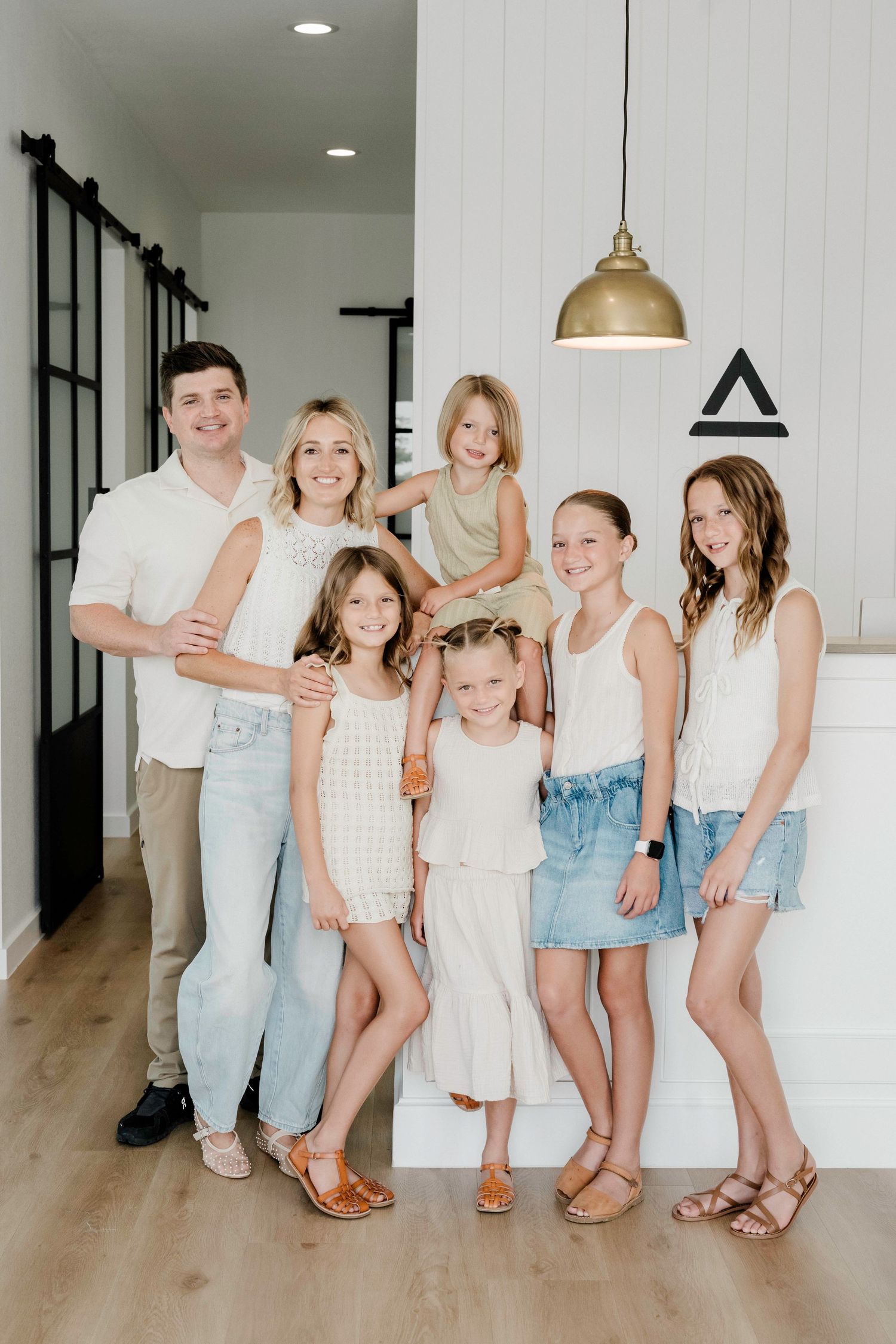 A family in coordinated white and light blue outfits poses together in a modern interior with hardwood floors and brass lighting.