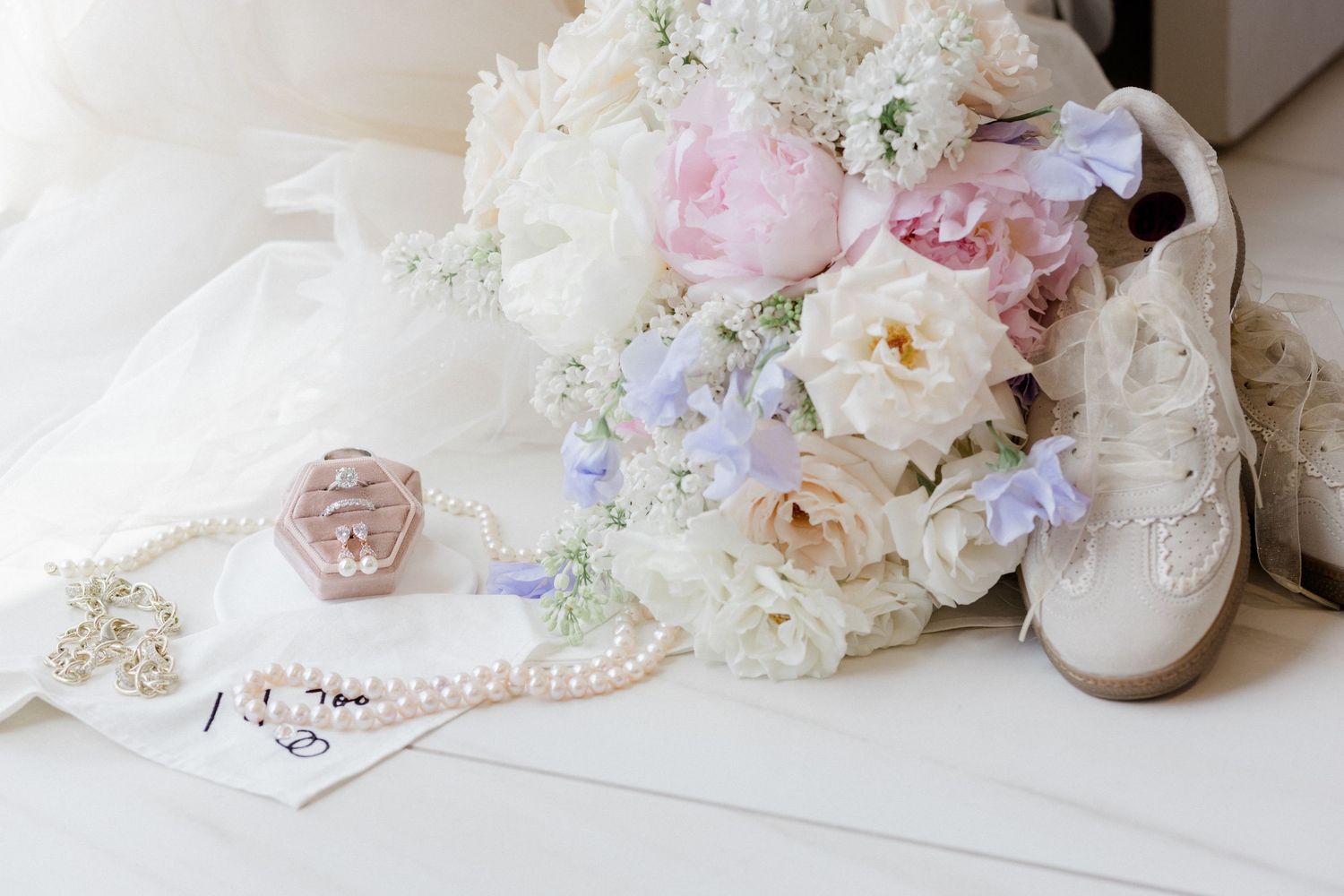 Bridal flat lay with pink peony bouquet, pearl necklace, ring box, earrings, and white sneakers on a white surface.