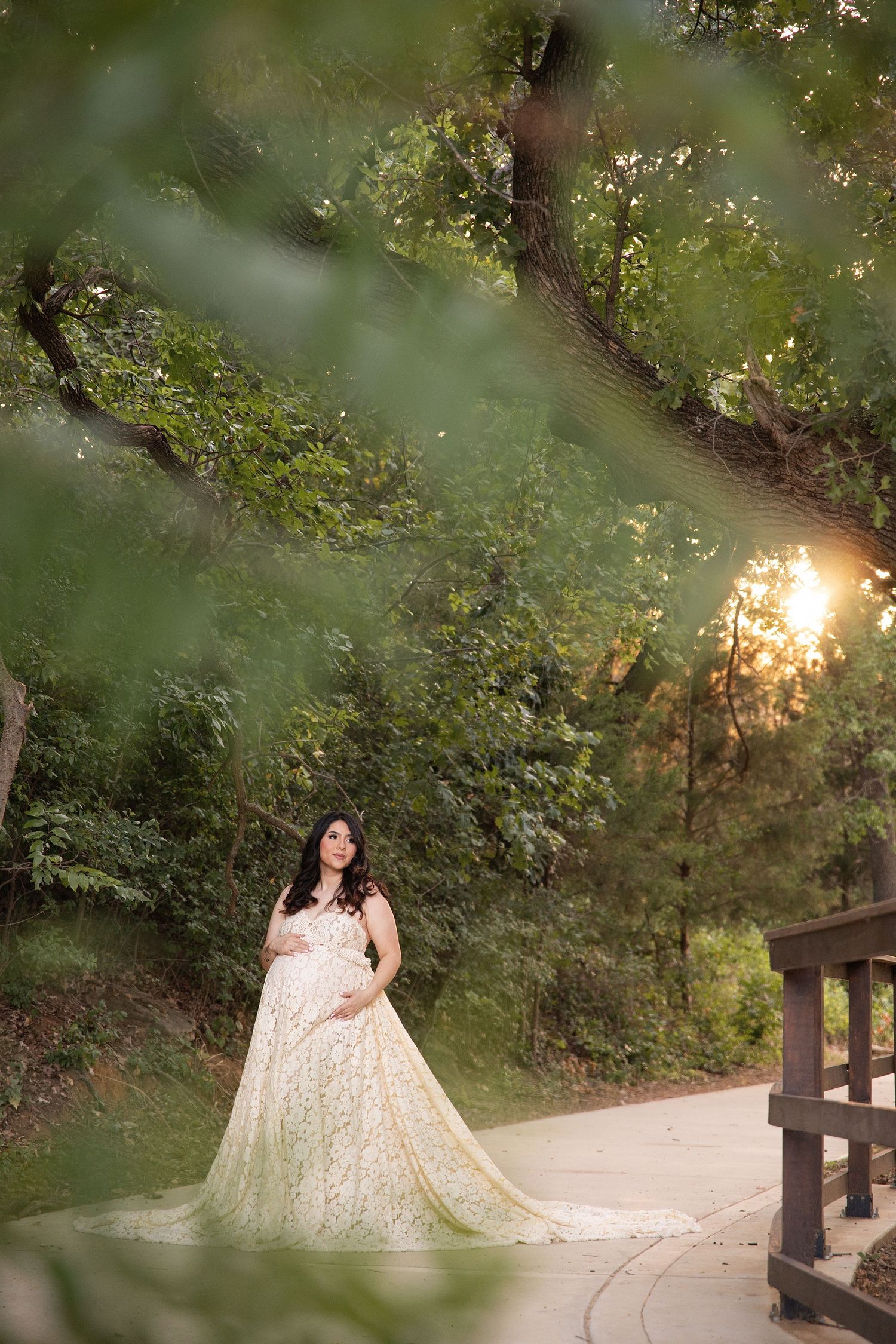 Pregnant woman in elegant white lace gown poses on garden path surrounded by lush green trees at golden hour.