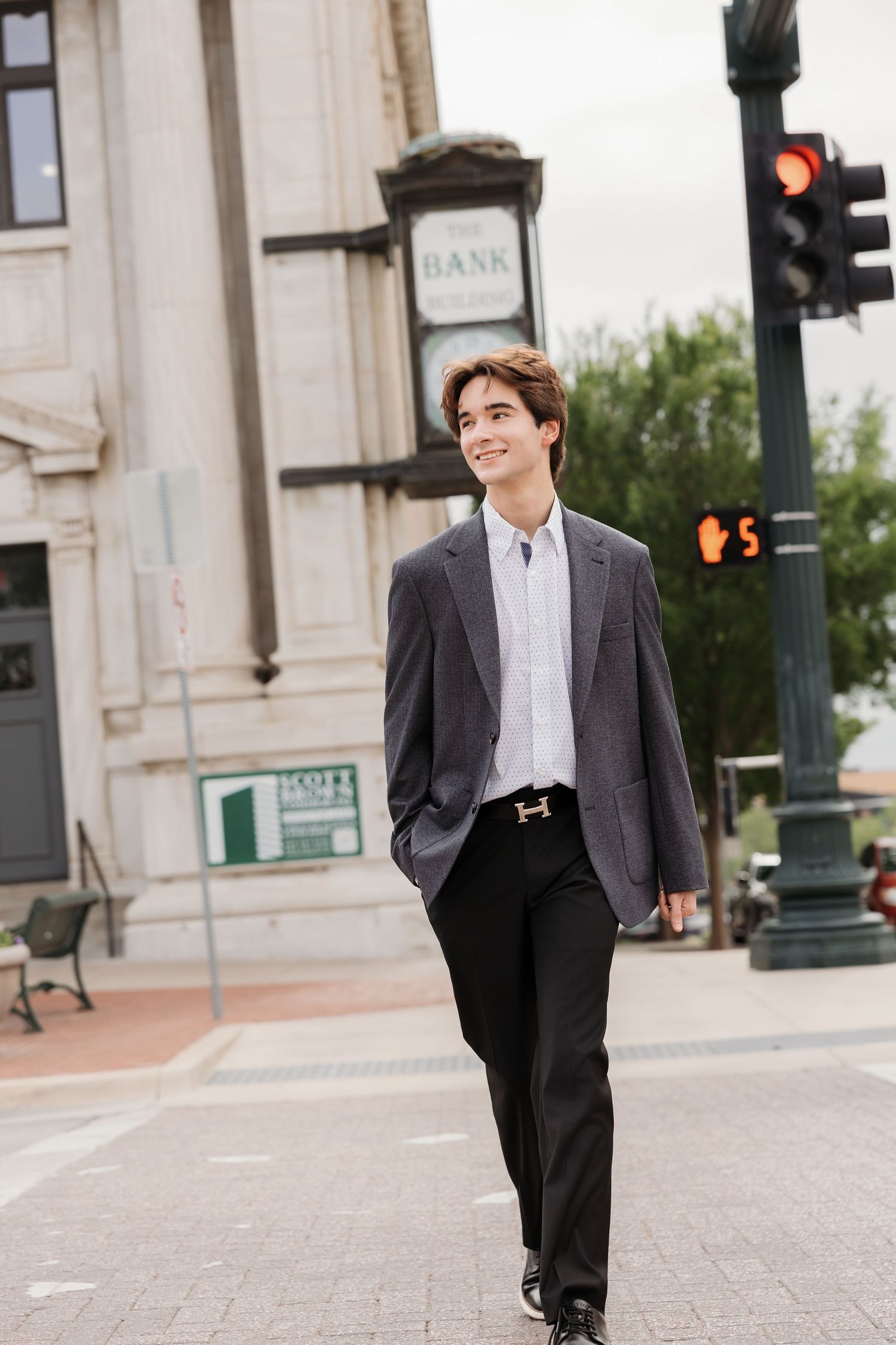 Young man in gray blazer and black pants walks confidently across a city street corner near a bank building.