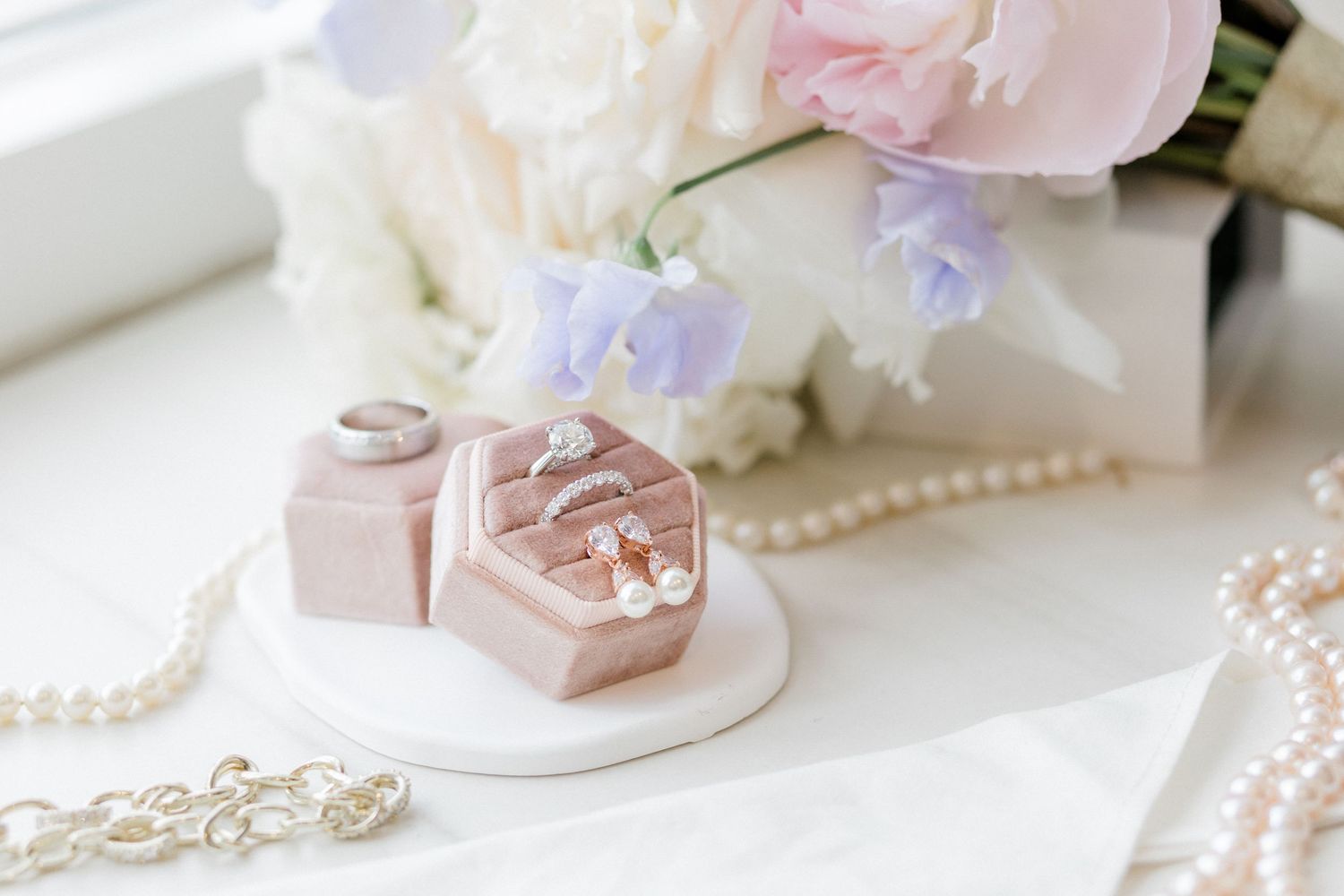 Elegant wedding flat lay with pink velvet ring box, pearl necklace, and pastel floral bouquet on white marble.