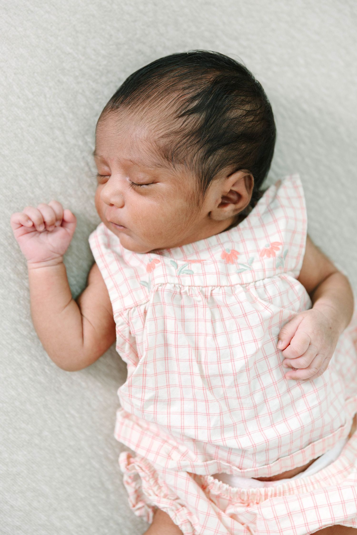 Newborn baby girl sleeping peacefully in a pink gingham dress with floral embroidery details.