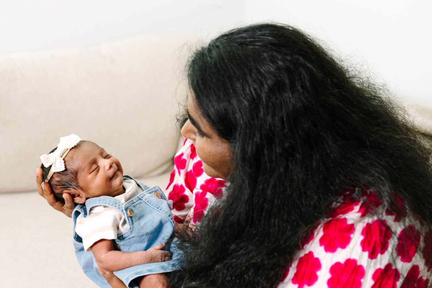 A woman in a red floral top tenderly holds a newborn baby wearing a denim outfit and white bow headband.