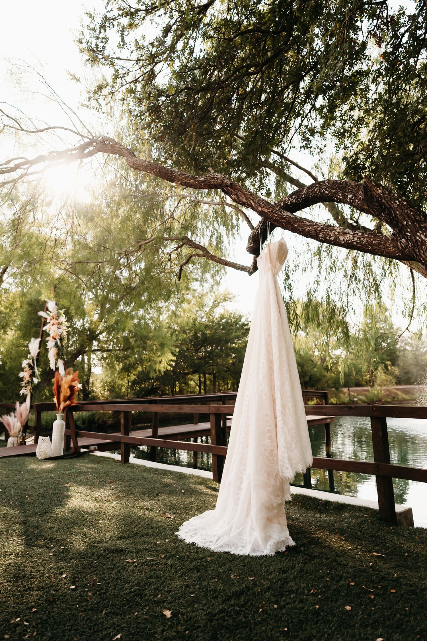 A white lace wedding dress hangs elegantly from a tree branch at sunset, creating a romantic outdoor setting.
