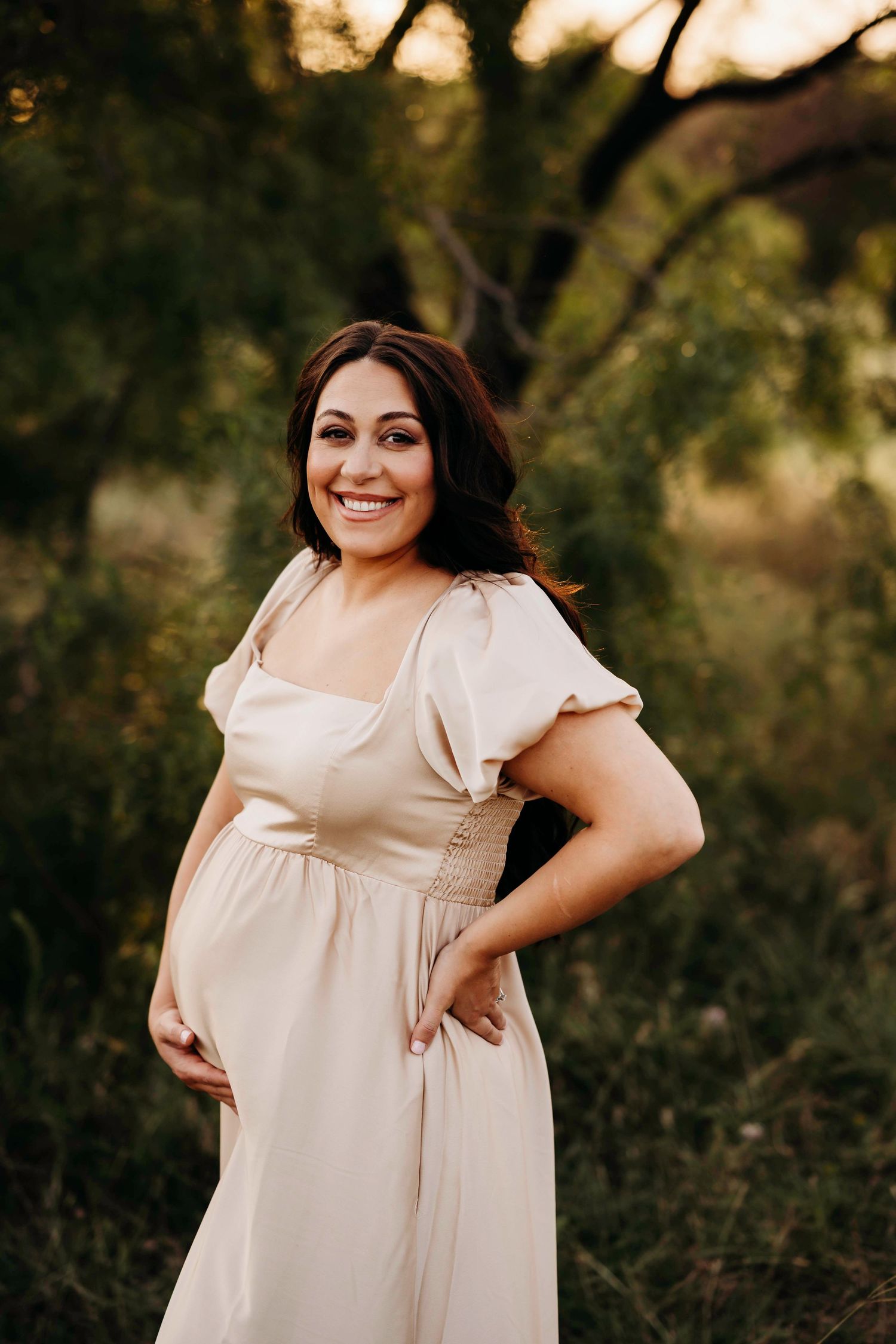 A woman in a cream-colored dress poses outdoors during golden hour in a natural setting.
