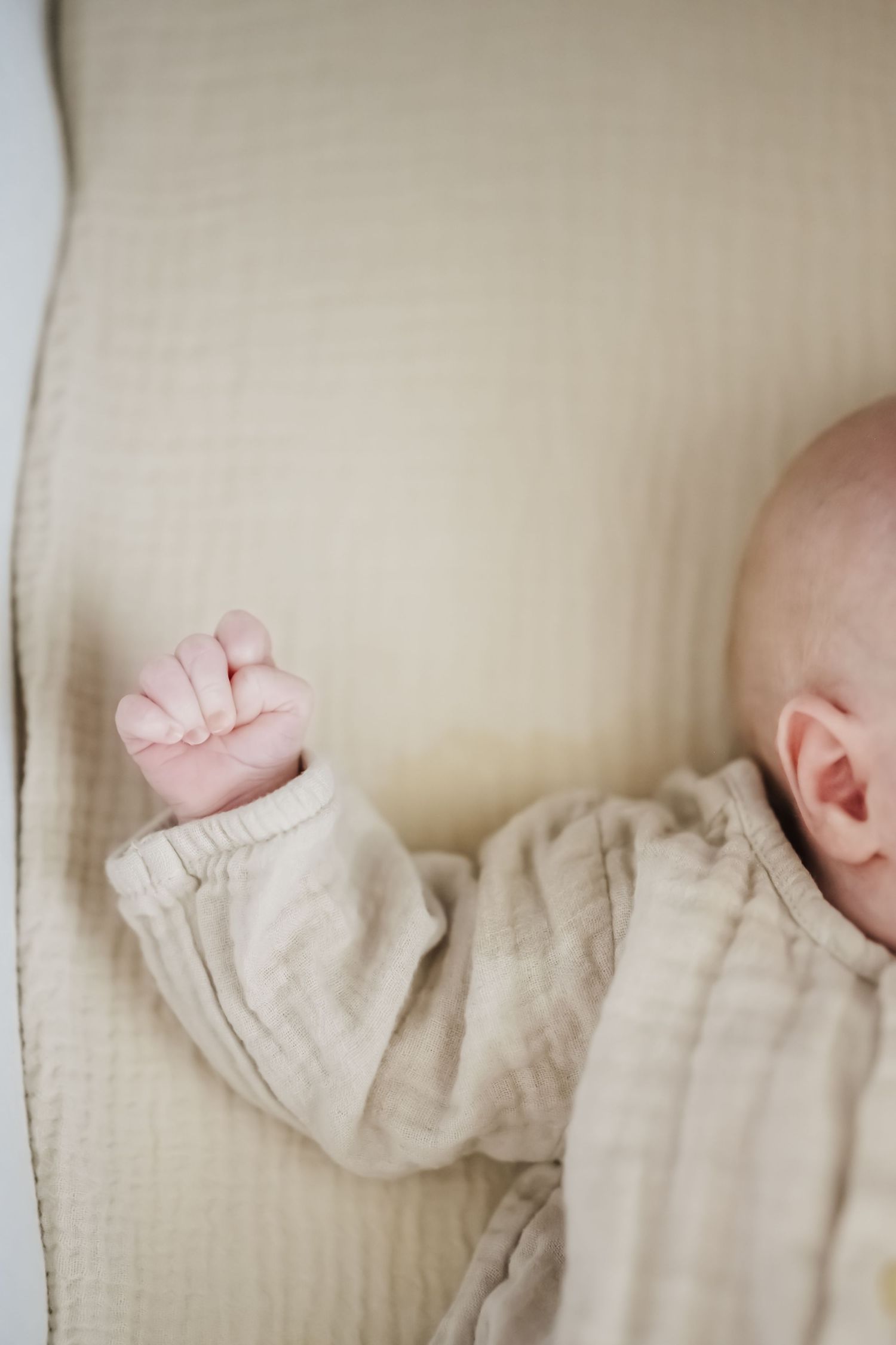 A baby wearing a beige linen outfit lies on a cream-colored surface.