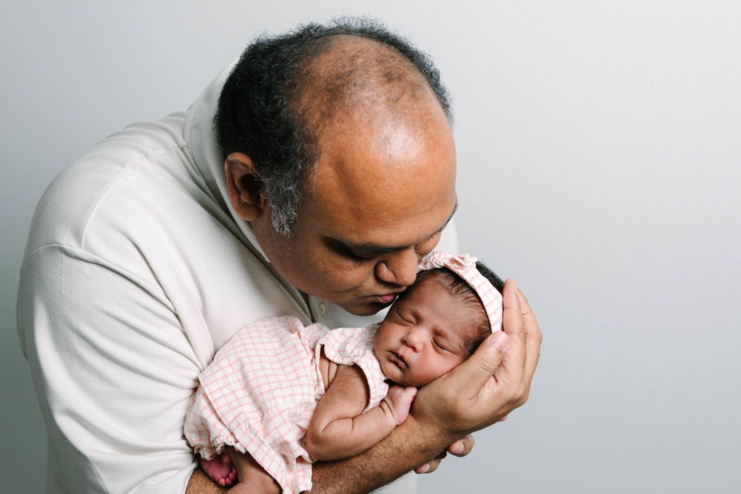 A bald man tenderly kisses a sleeping newborn baby dressed in pink, cradling her gently in his arms.